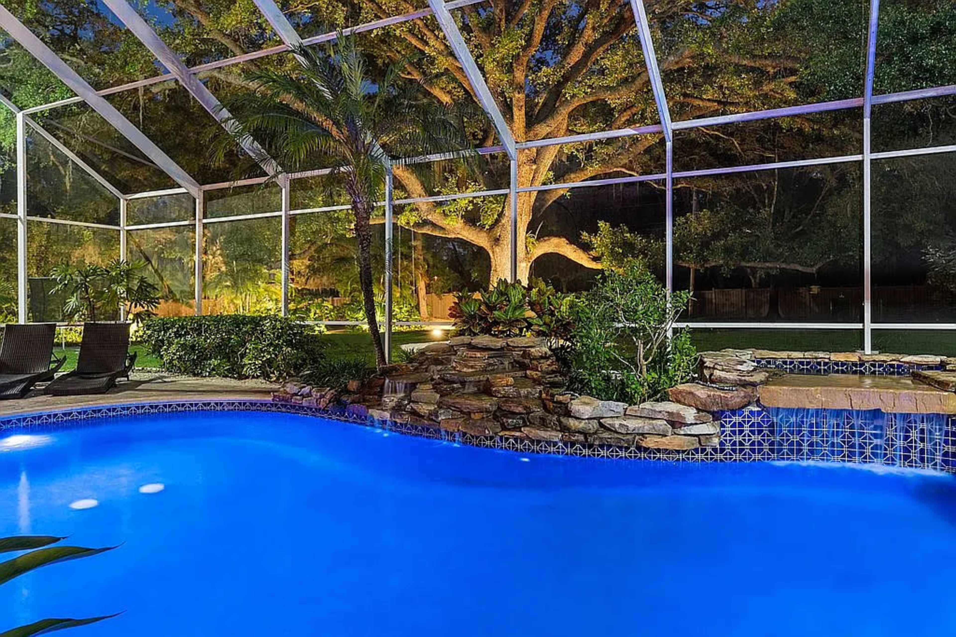 The image shows a backyard swimming pool with a rock waterfall, surrounded by lush greenery and a screened enclosure, illuminated at night.