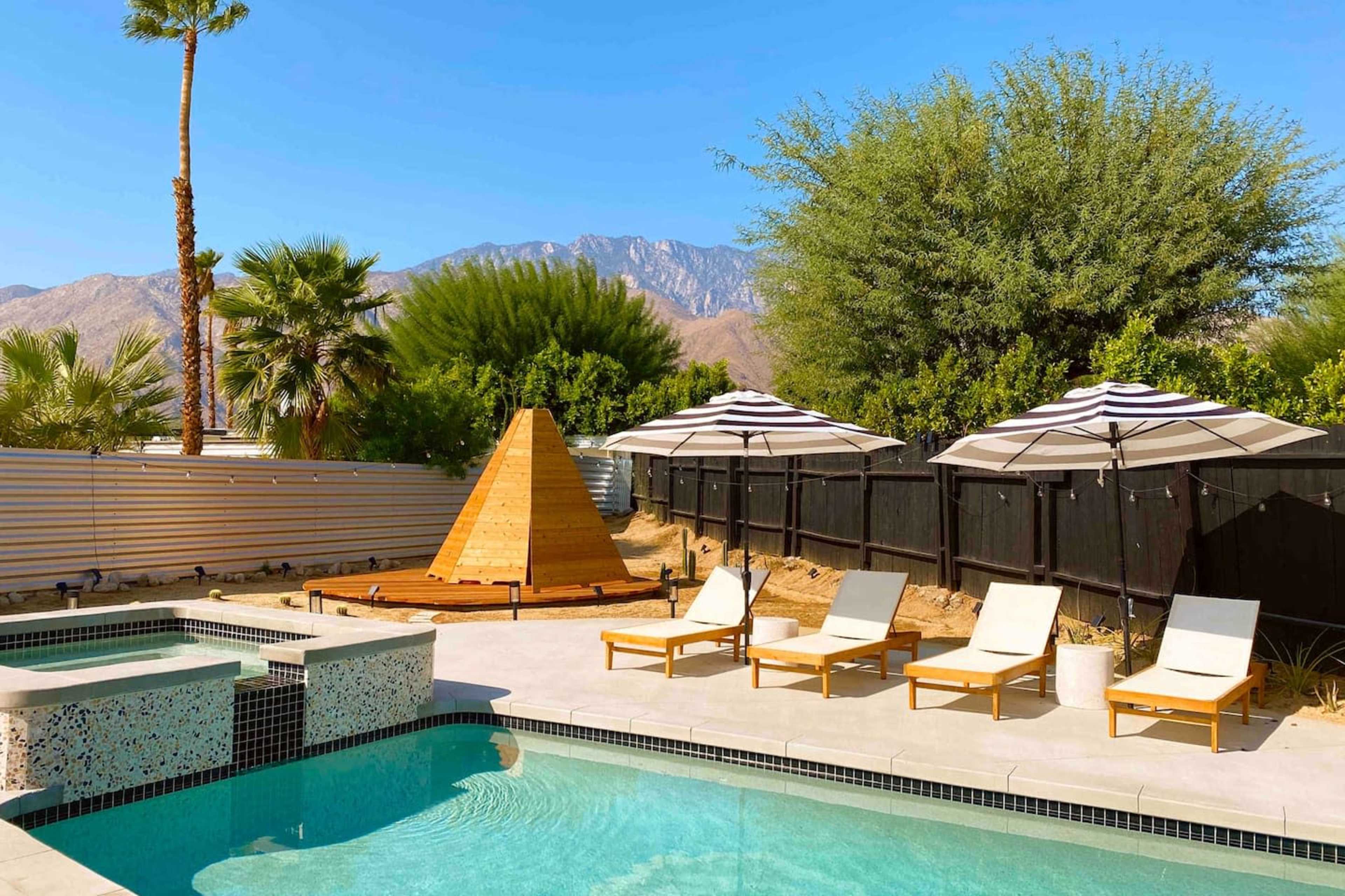 The image shows a swimming pool surrounded by lounge chairs and umbrellas, with a wooden pyramid structure and mountains in the background.