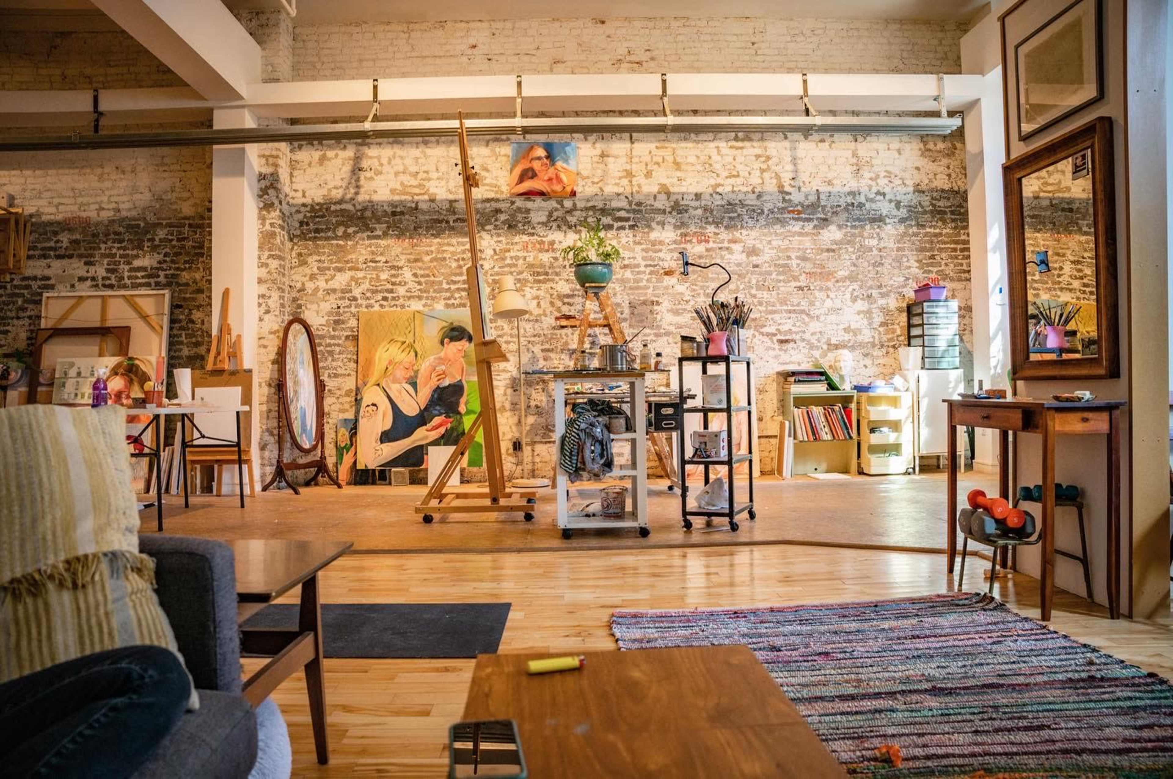 An artist's studio features wooden floors, exposed brick walls, an easel with artwork, various art supplies, and a cozy seating area.