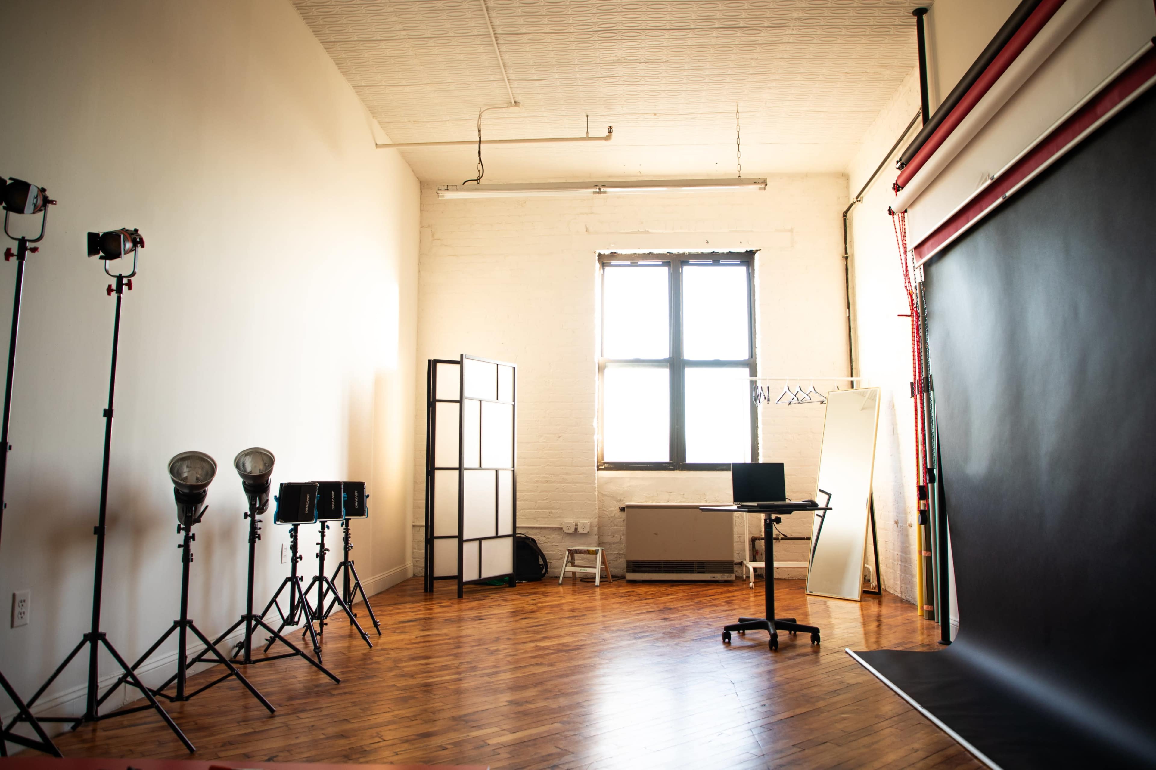 A photography studio features a large window, a backdrop setup, lighting equipment on stands, and a table with a computer.