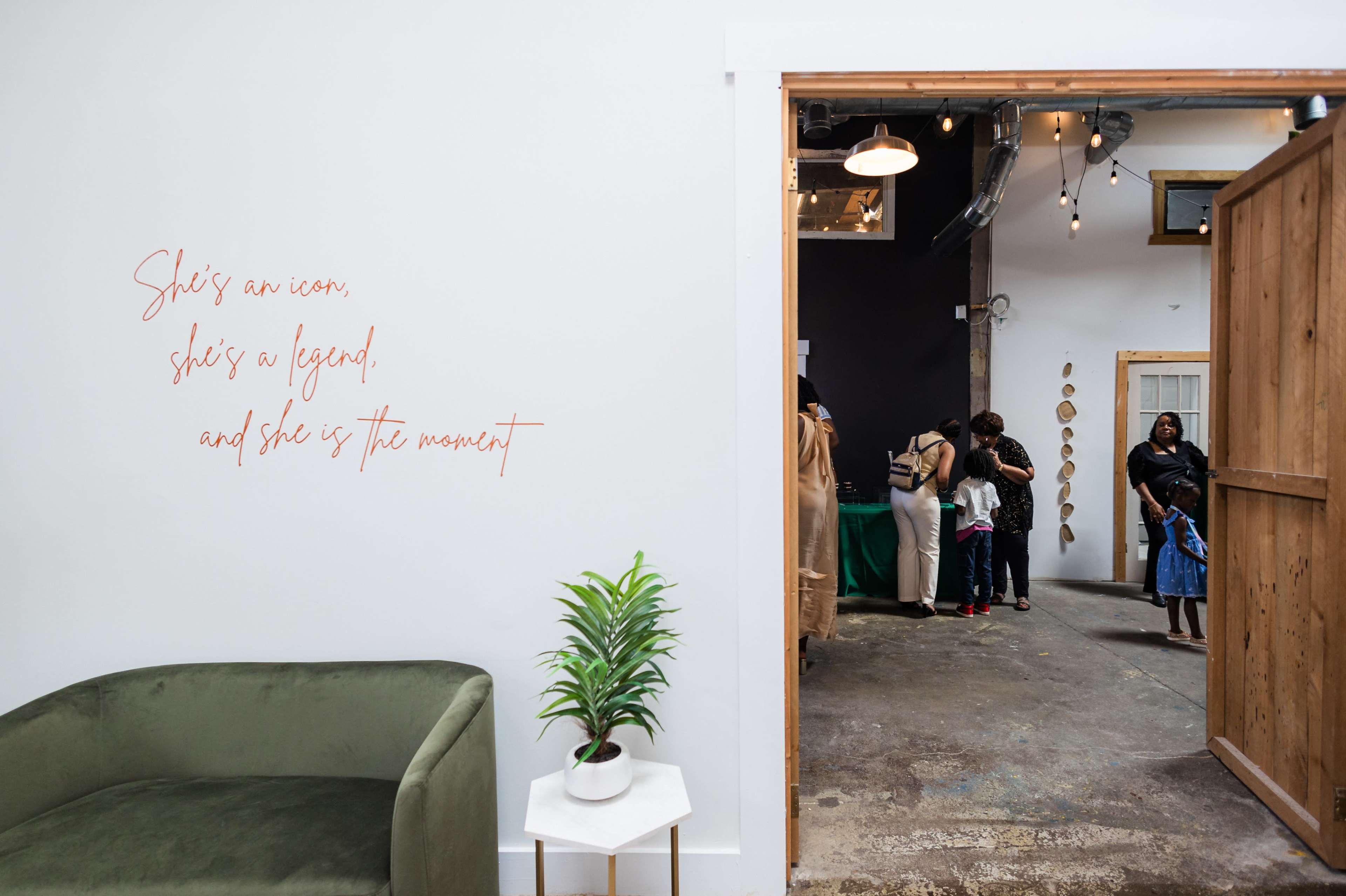 The image shows an interior space with an open wooden door leading to a gathering of people, with a pink script quote on the wall stating, "She's an icon, she's a legend, and she is the moment."