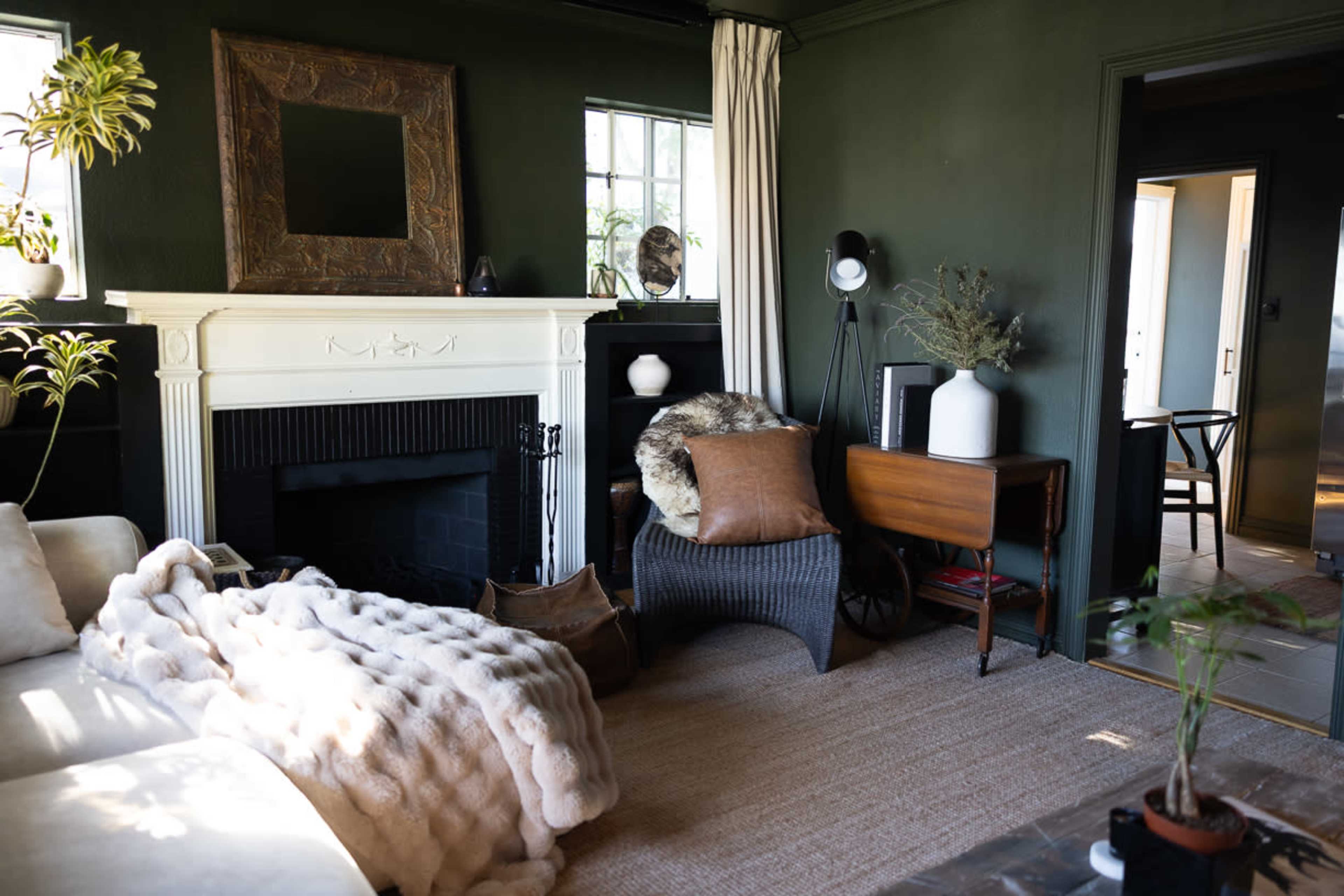 A cozy living room features a white fireplace, dark green walls, a plush couch, and various decorative elements including plants and art.