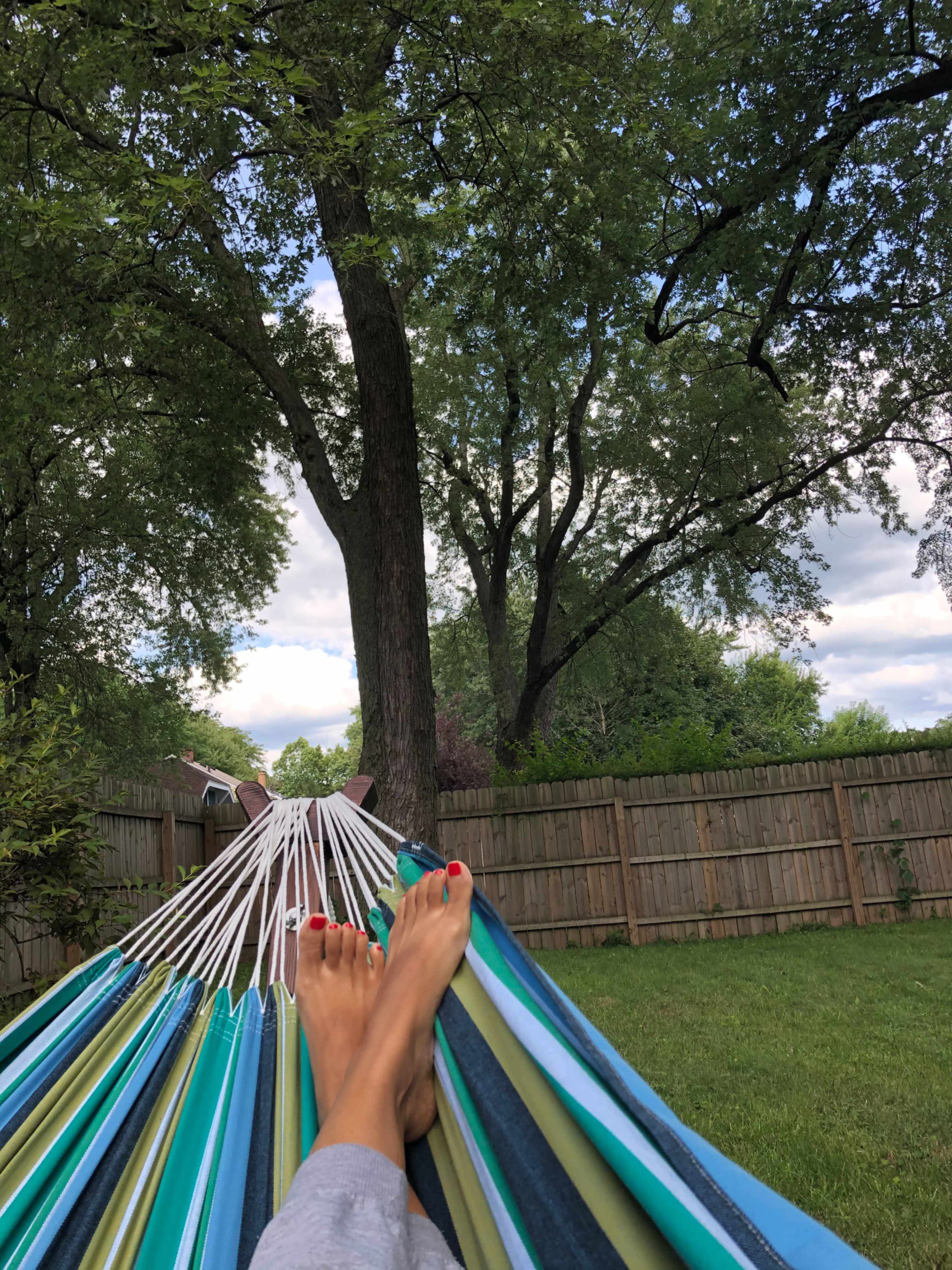A person relaxes on a colorful hammock in a backyard, with trees and a wooden fence in the background.