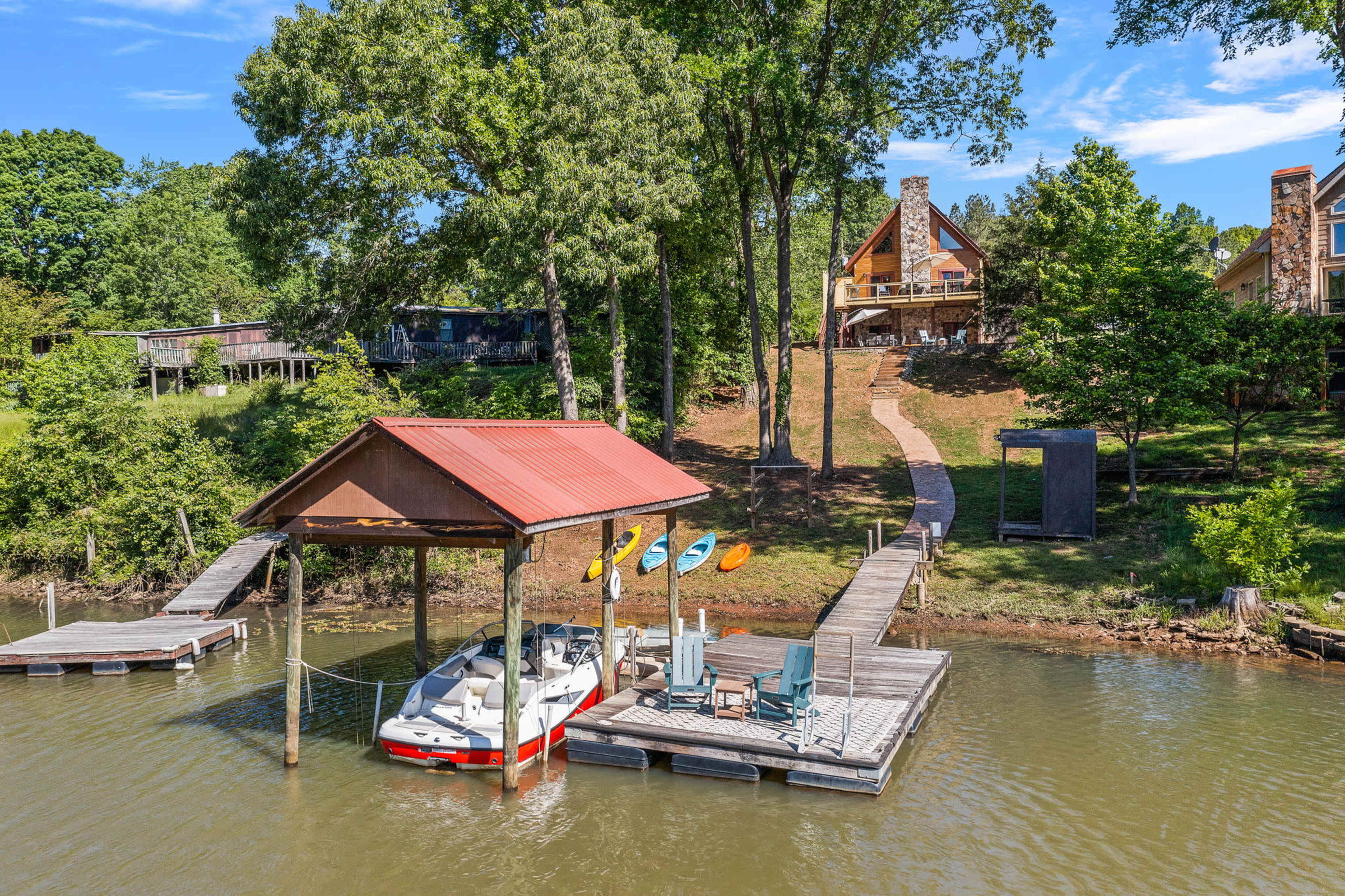 A boat dock with a red-roofed shelter is located on a calm waterway, surrounded by trees and a pathway leading to a house on a hillside.