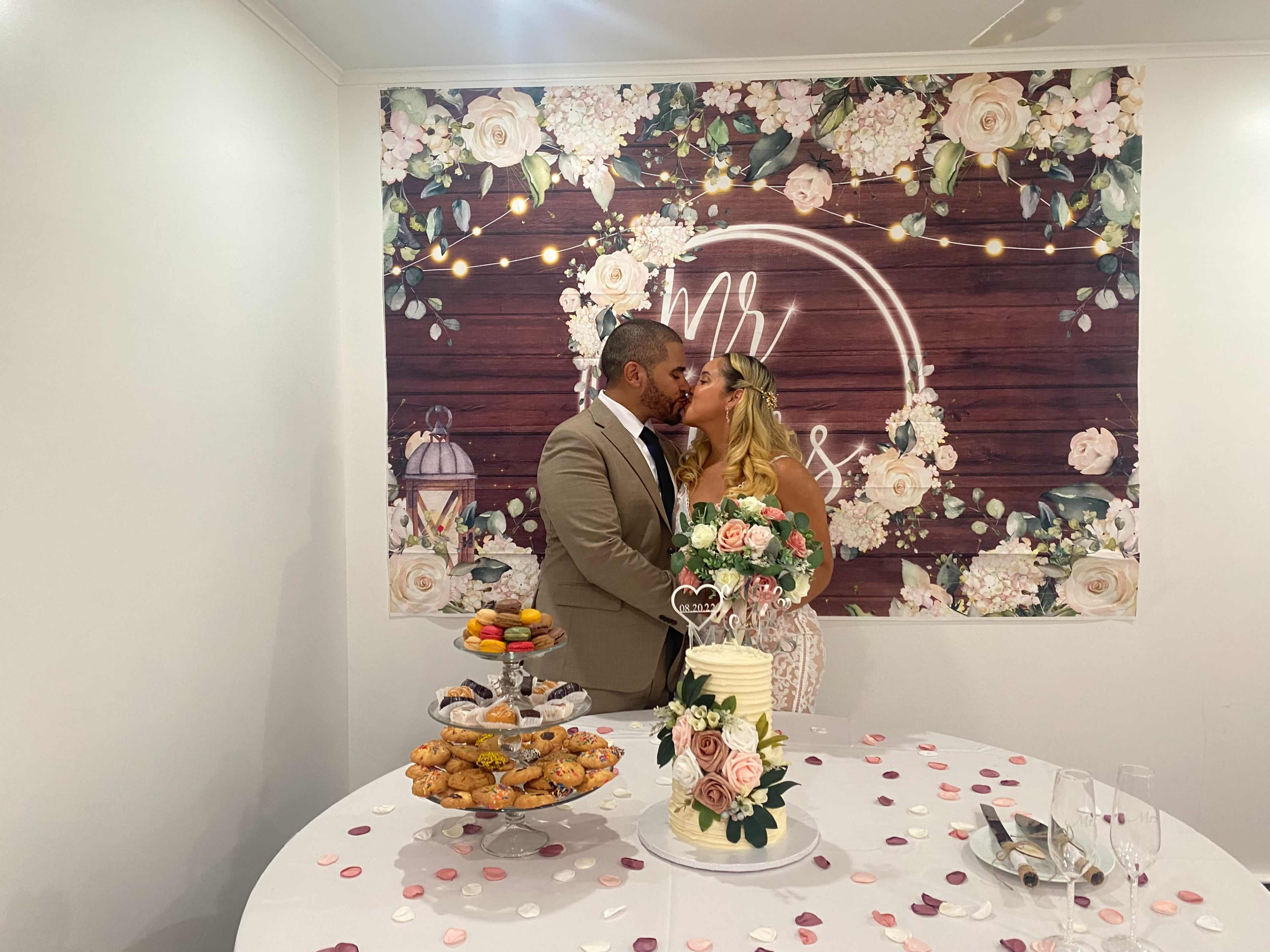 A couple kisses in front of a floral backdrop while standing beside a table adorned with sweets and a wedding cake.