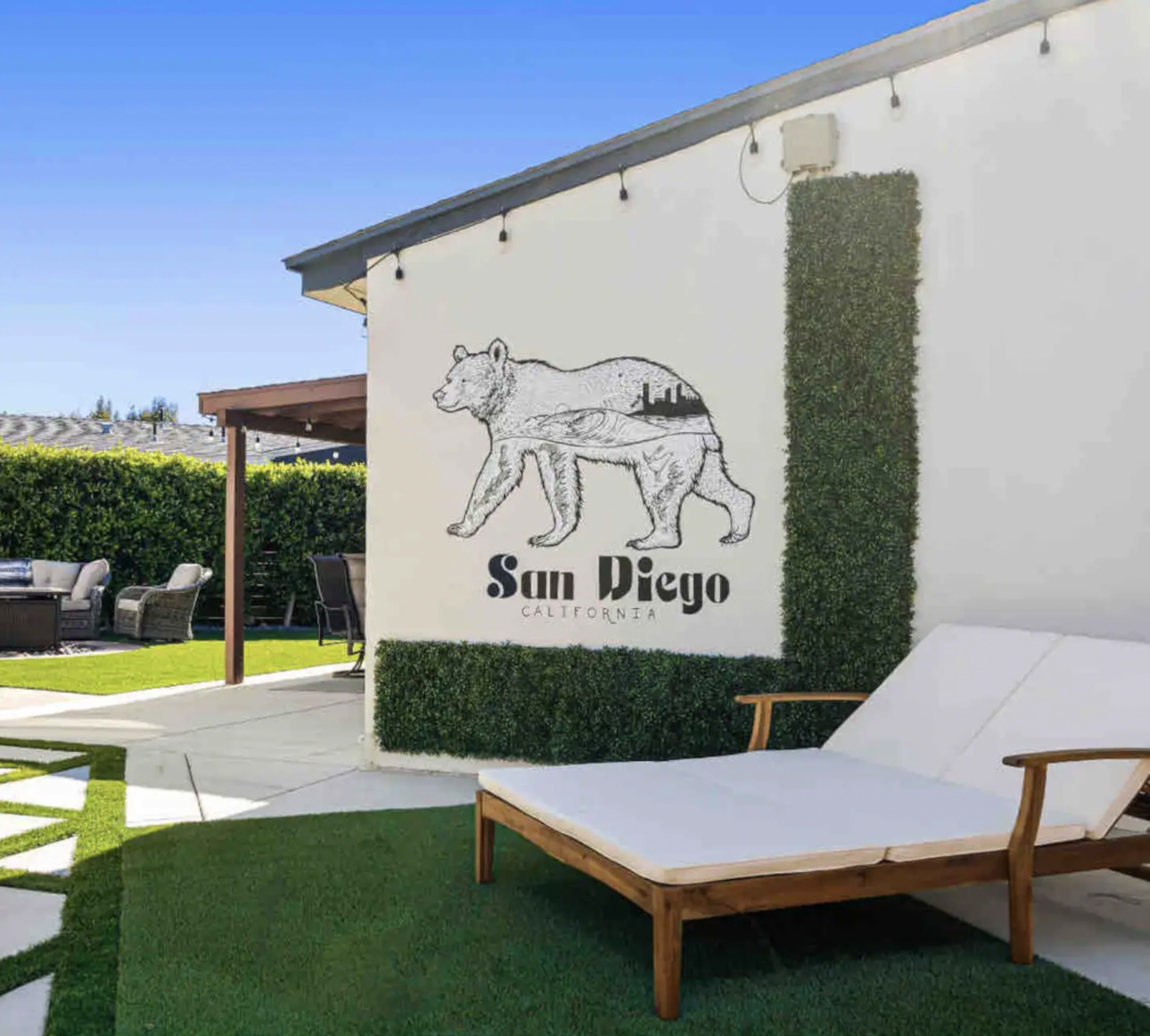 A wooden lounge chair sits on green grass beside a wall featuring a bear silhouette filled with a city skyline and the text "San Diego, California."