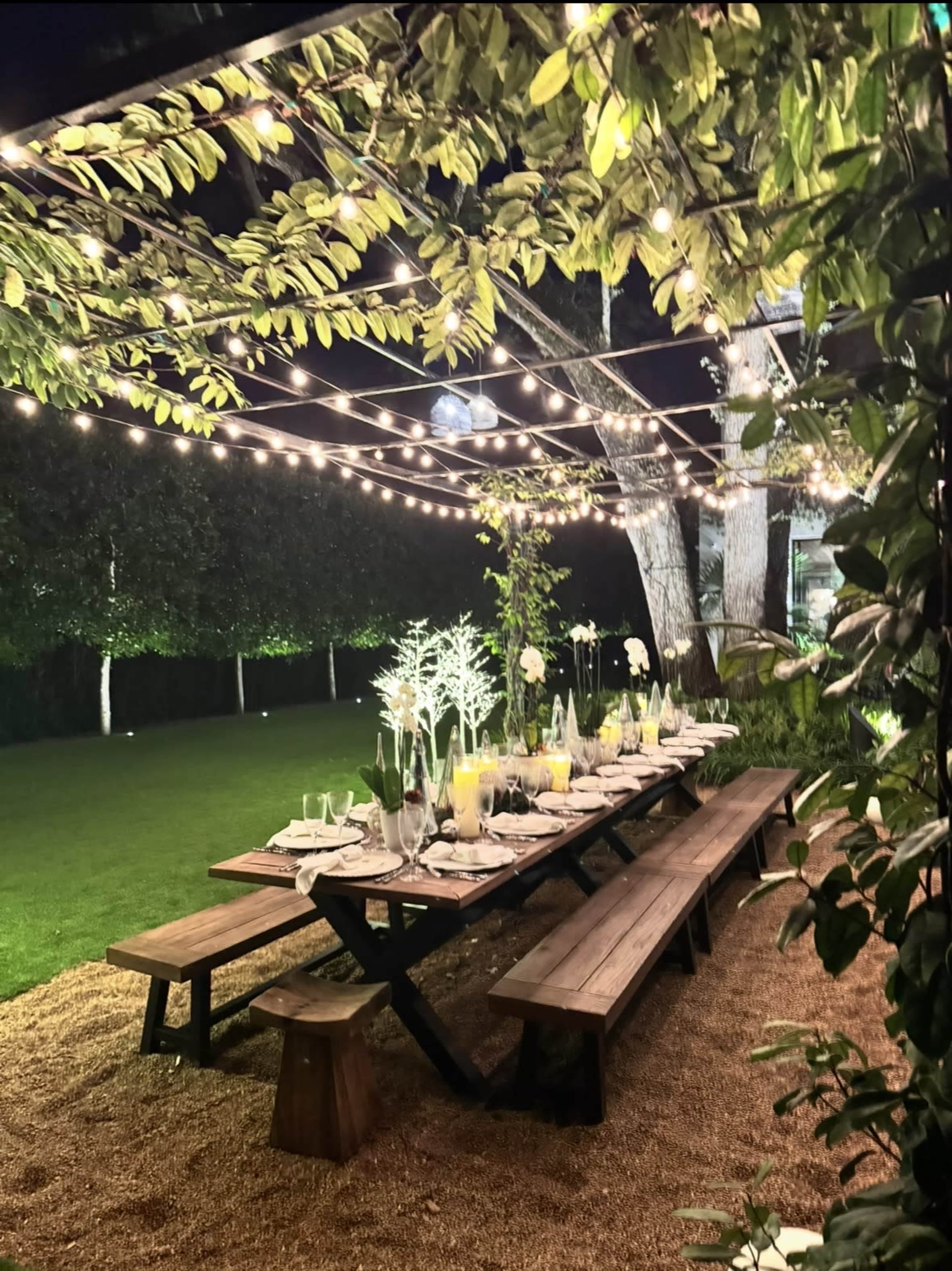 A long wooden table is set for dinner under string lights and surrounded by greenery in a garden at night.