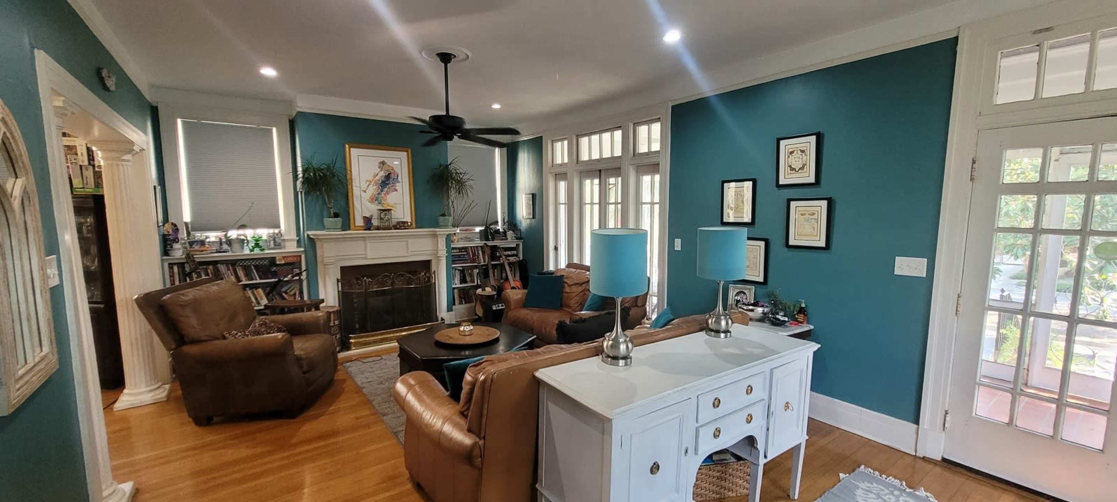 The image shows a living room with teal walls, a fireplace framed by bookshelves, and two brown armchairs opposite a white console table with blue lamps.