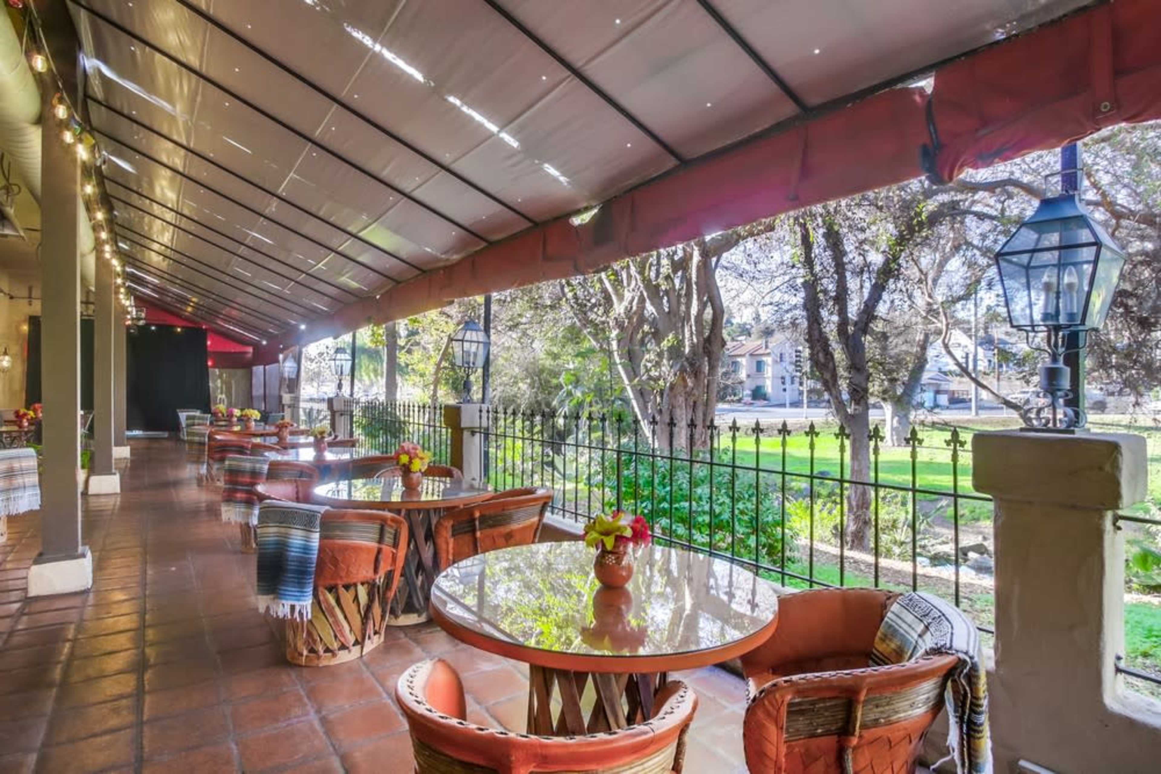 A patio area with several round tables and chairs is lined with plants and has a view of a grassy landscape.