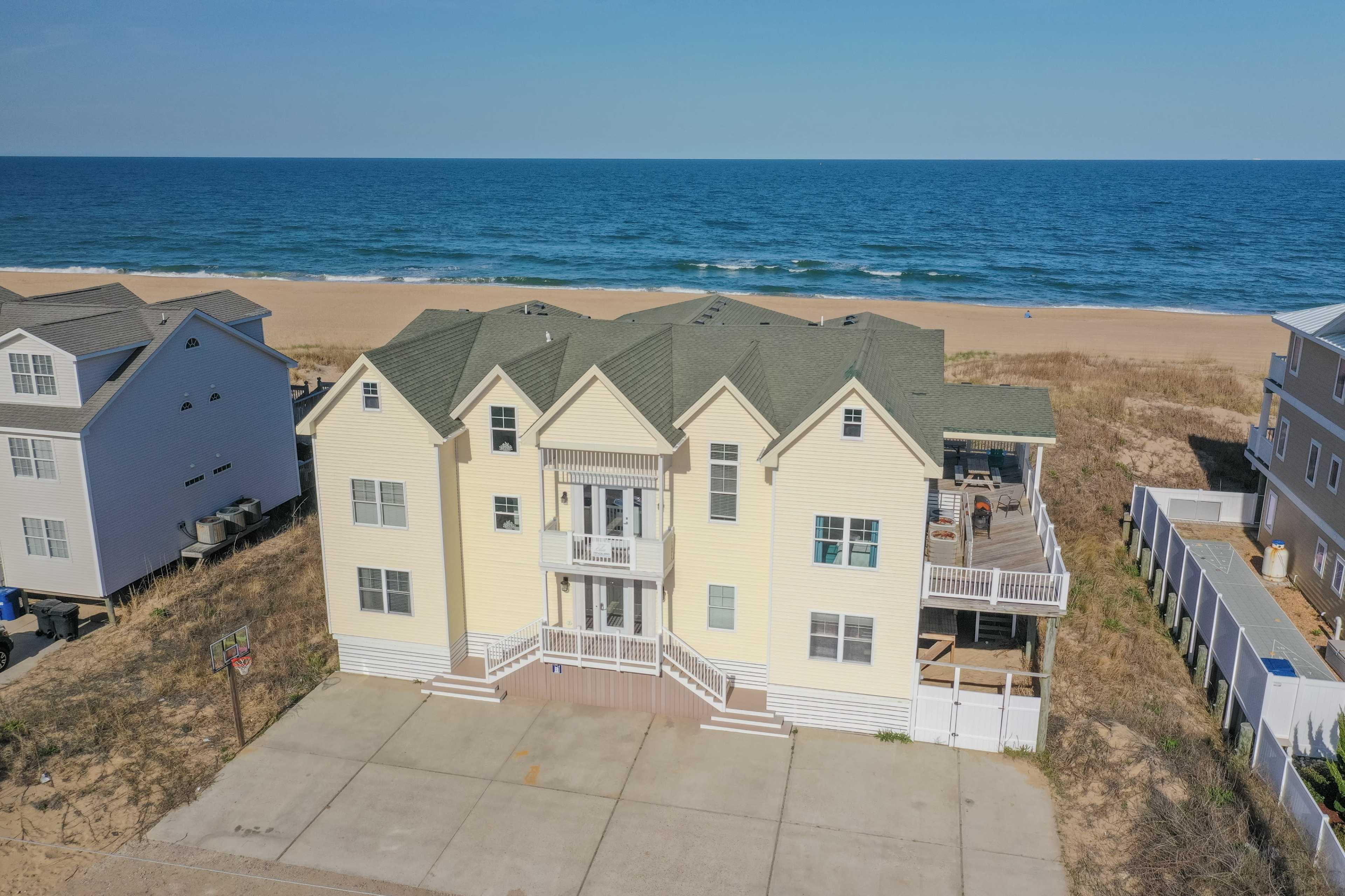 A large yellow beach house with multiple gabled roofs is situated near a sandy shoreline and the ocean.