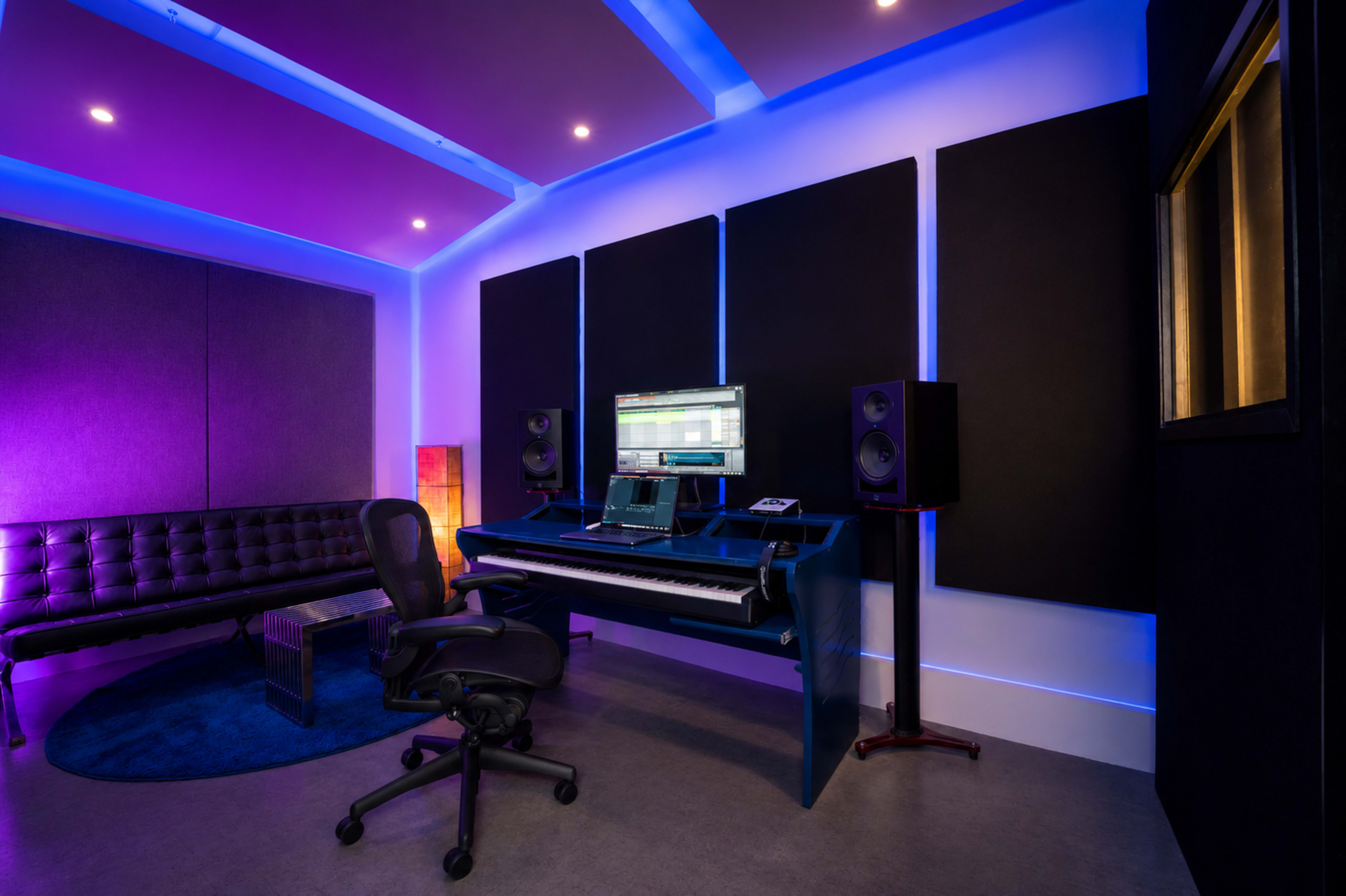 The image shows a modern music studio featuring a digital keyboard, computer setup, and soundproof wall panels illuminated by colorful lighting.