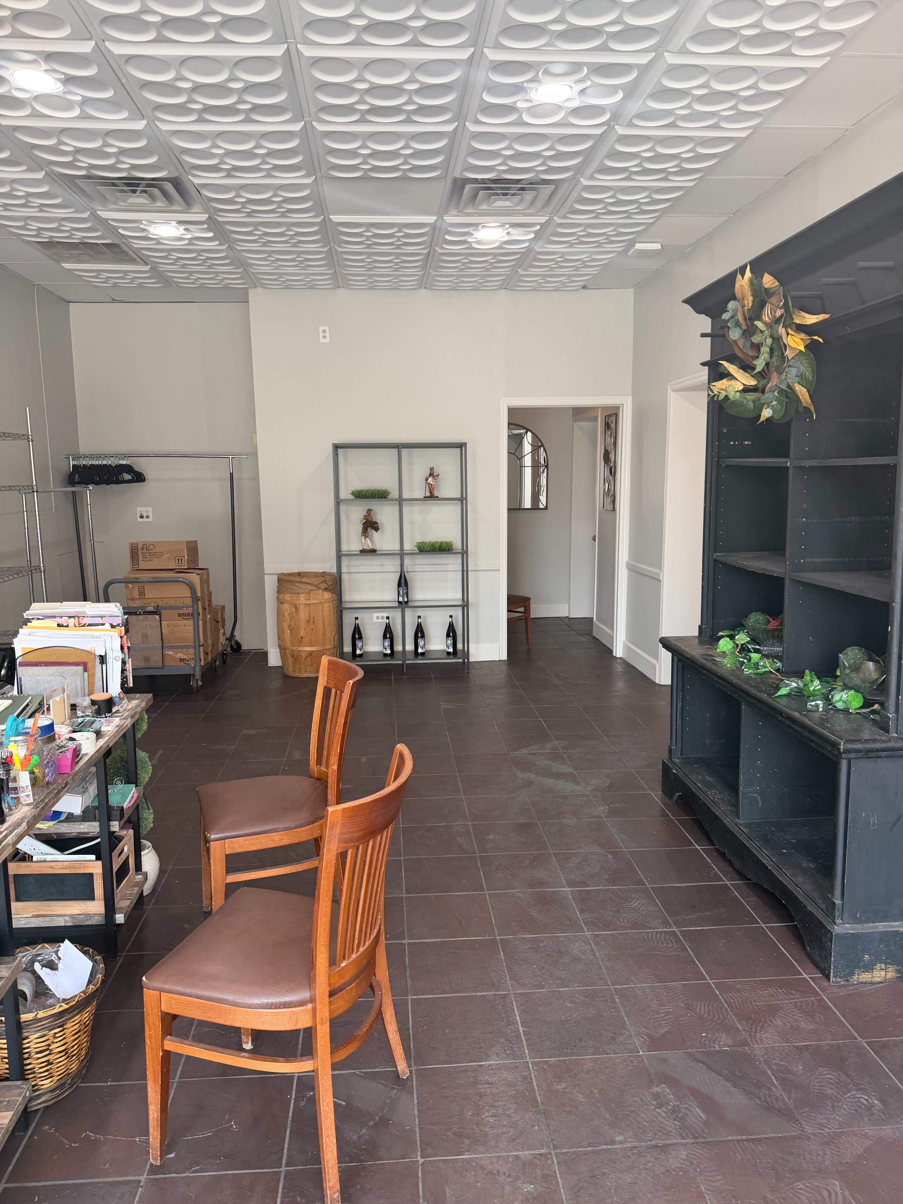 The image shows a vacant interior space with two wooden chairs, a shelf with decorative items, and a tiled floor.