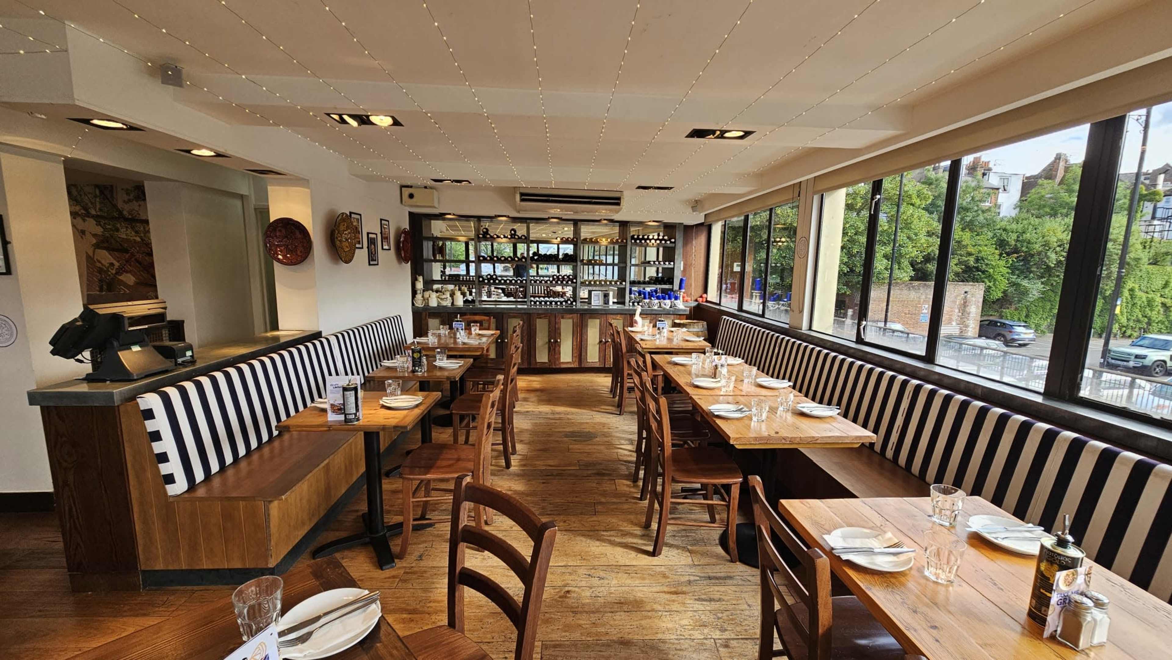 The image shows a spacious restaurant interior with wooden tables, striped seating, and large windows letting in natural light.
