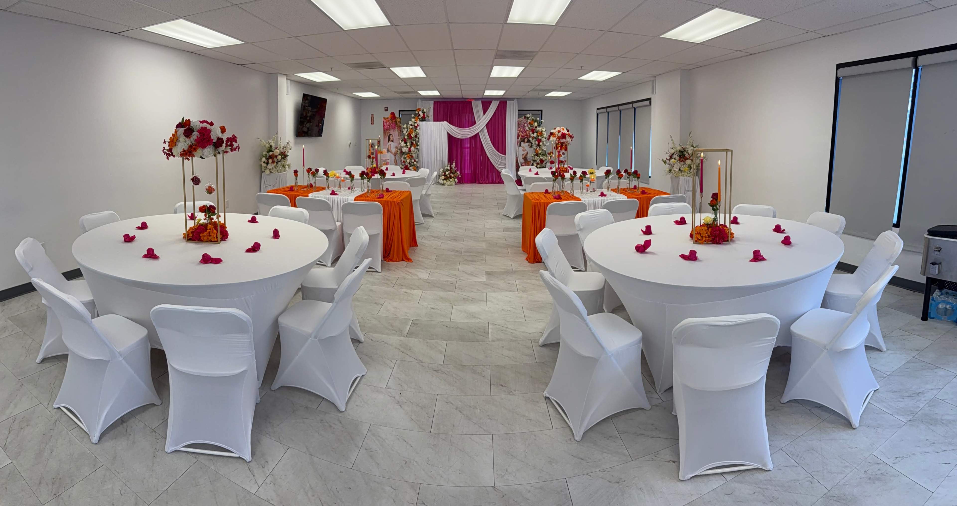 The image shows a banquet hall set up with round tables draped in white tablecloths and orange accent decorations, surrounded by floral arrangements and a backdrop featuring pink drapery.
