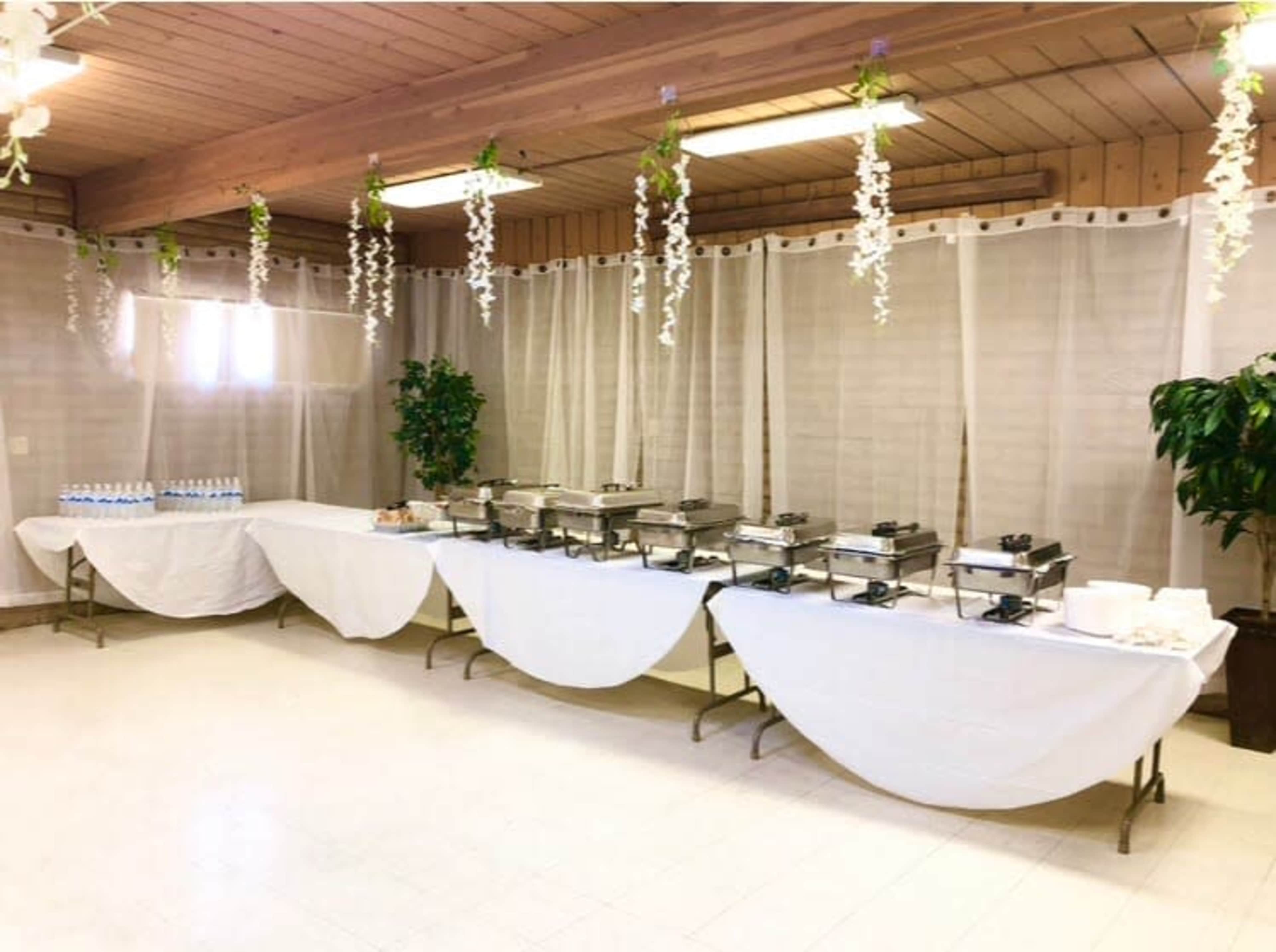 A buffet setup featuring multiple chafing dishes and tables covered with white tablecloths in a room with light-colored walls and hanging decorations.