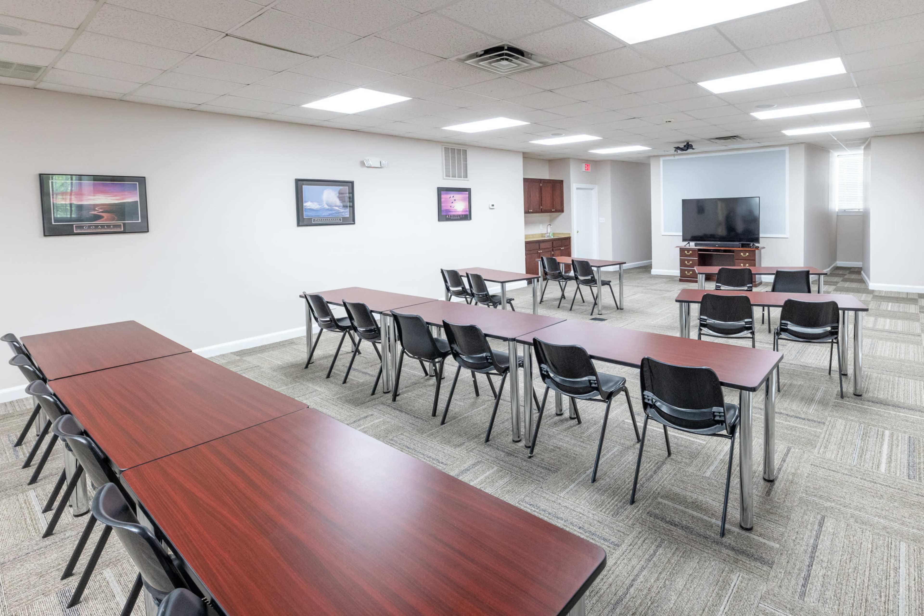 A meeting room with multiple tables arranged in rows and black chairs, along with a television and some framed artwork on the walls.