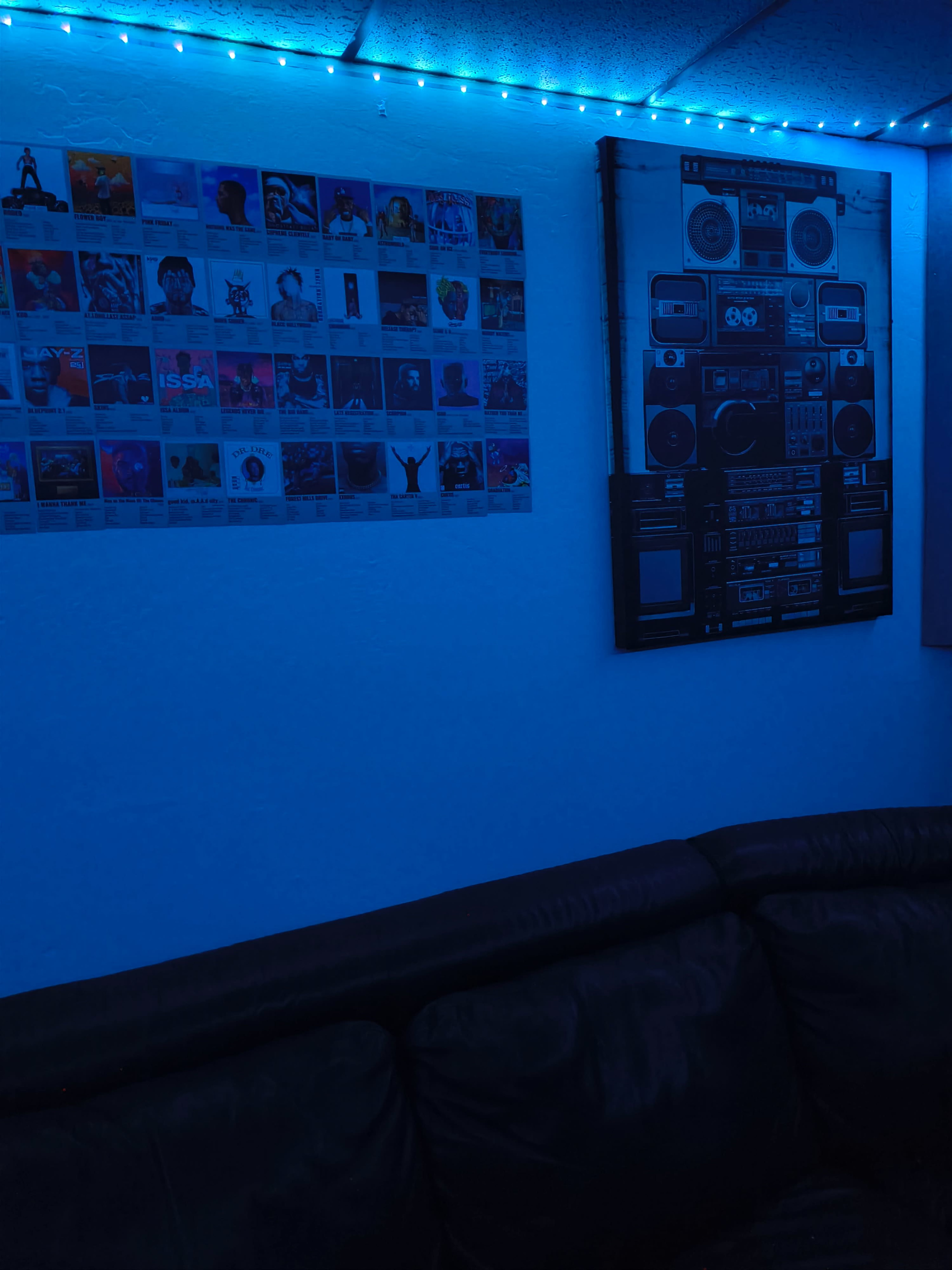 The image features a dimly lit room with blue ambient lighting, showcasing a wall adorned with a collage of album covers and a large artistic representation of a vintage boombox above a dark-colored couch.