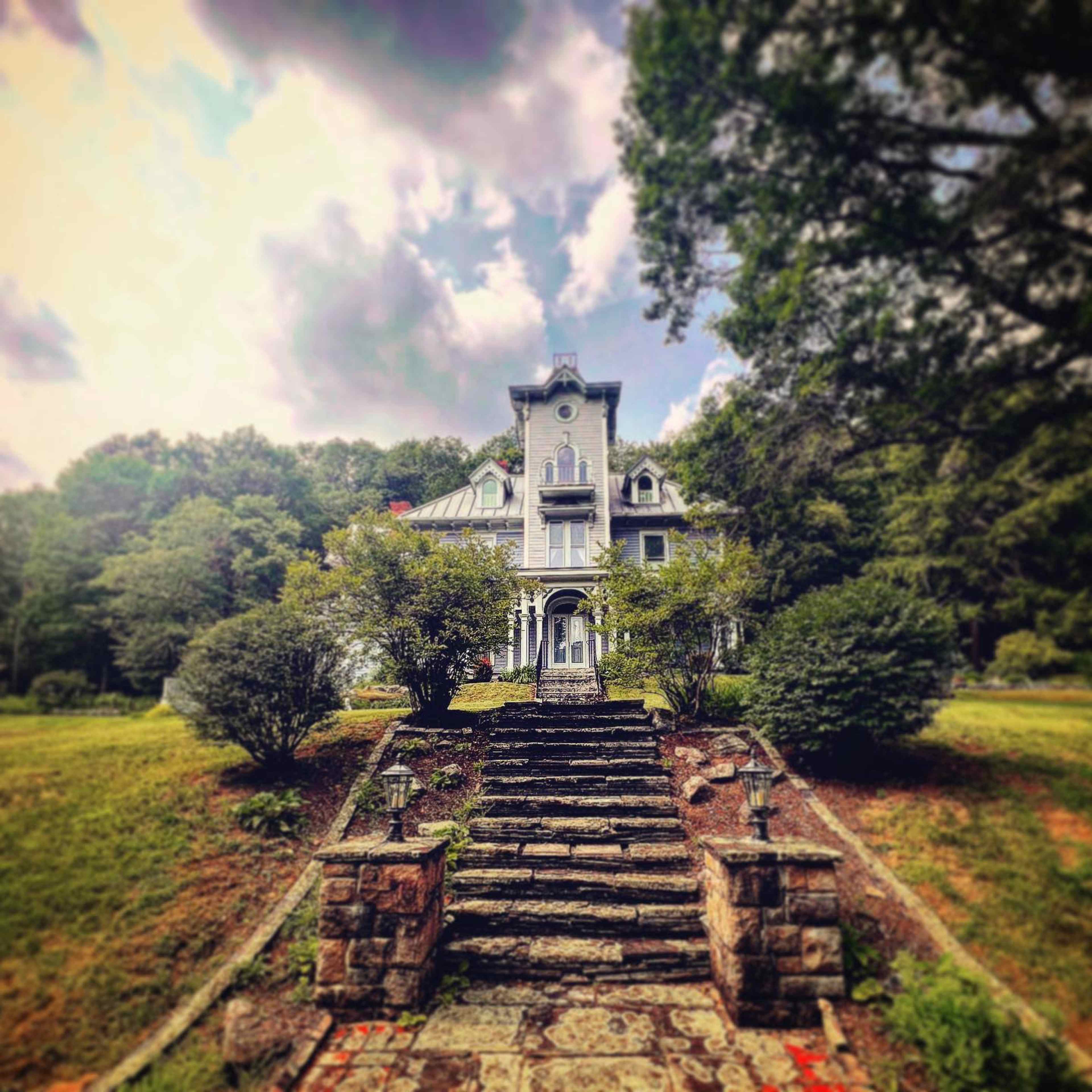 A large, two-story house with a tower and multiple gables is surrounded by well-kept gardens and a stone pathway.