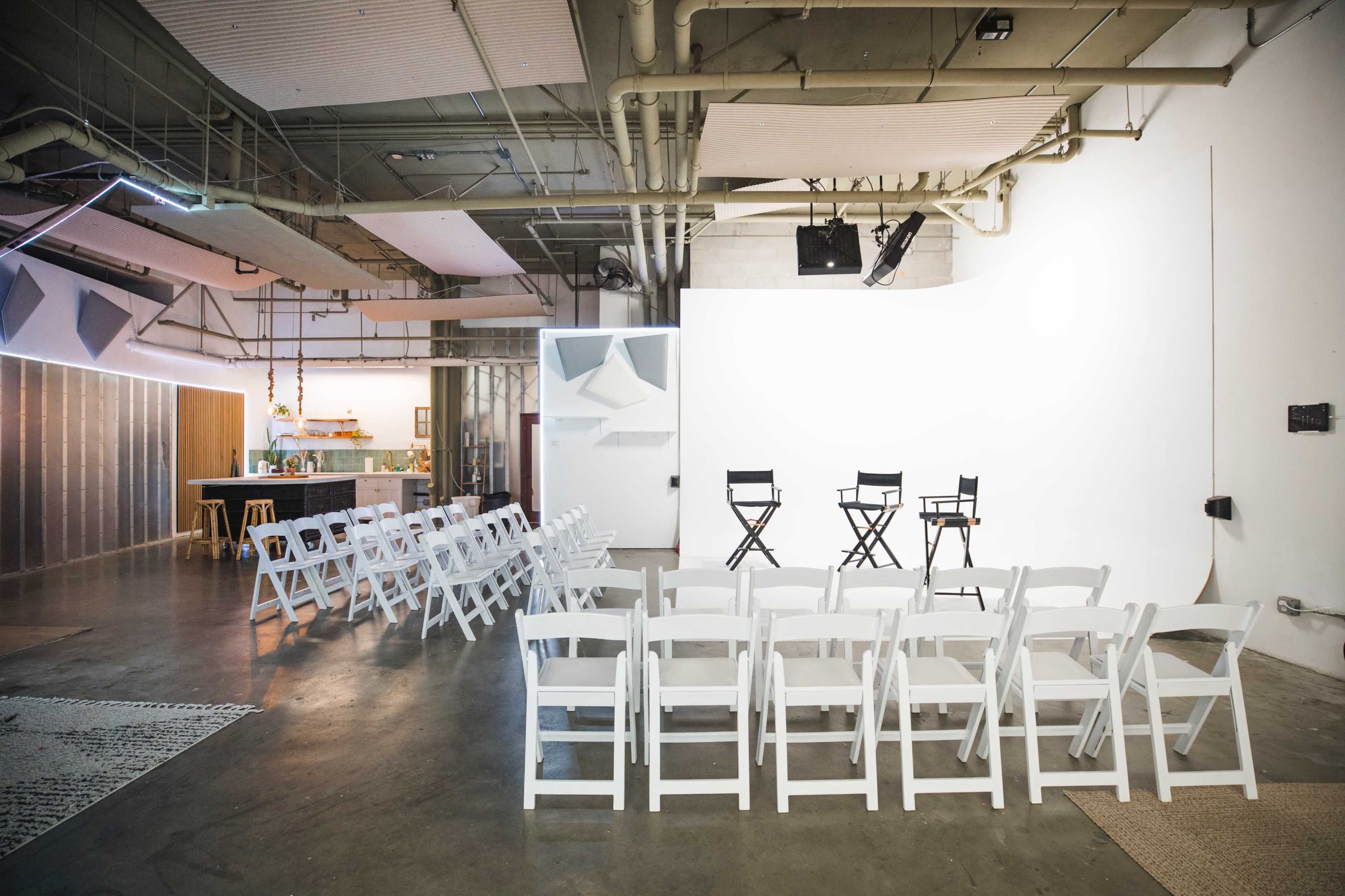 The image shows a minimalist studio setup with rows of white folding chairs facing a blank white screen, accompanied by a small kitchen area in the background.