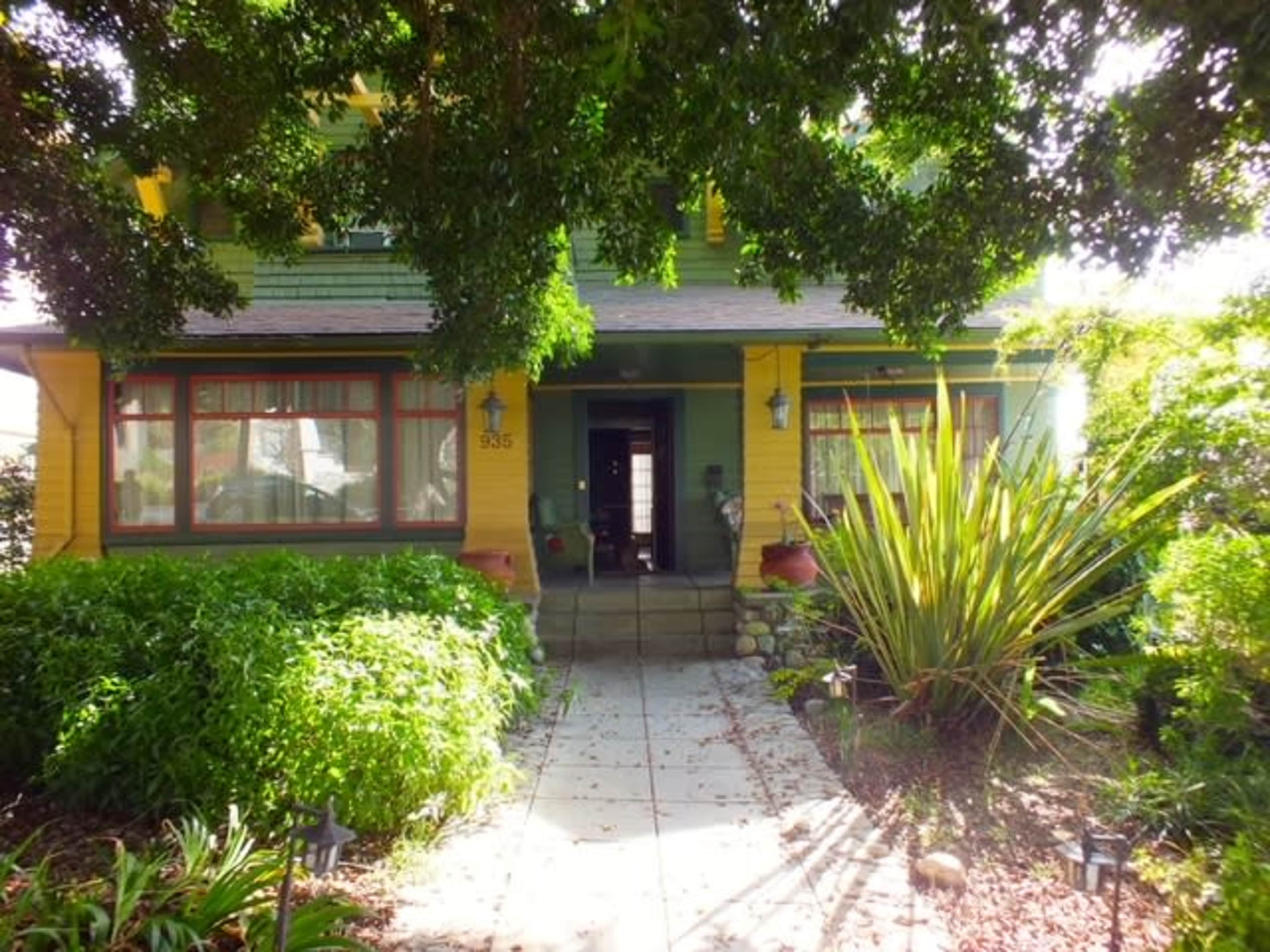 A two-story house with yellow and green exterior paint, surrounded by lush greenery and a stone pathway leading to the front door.
