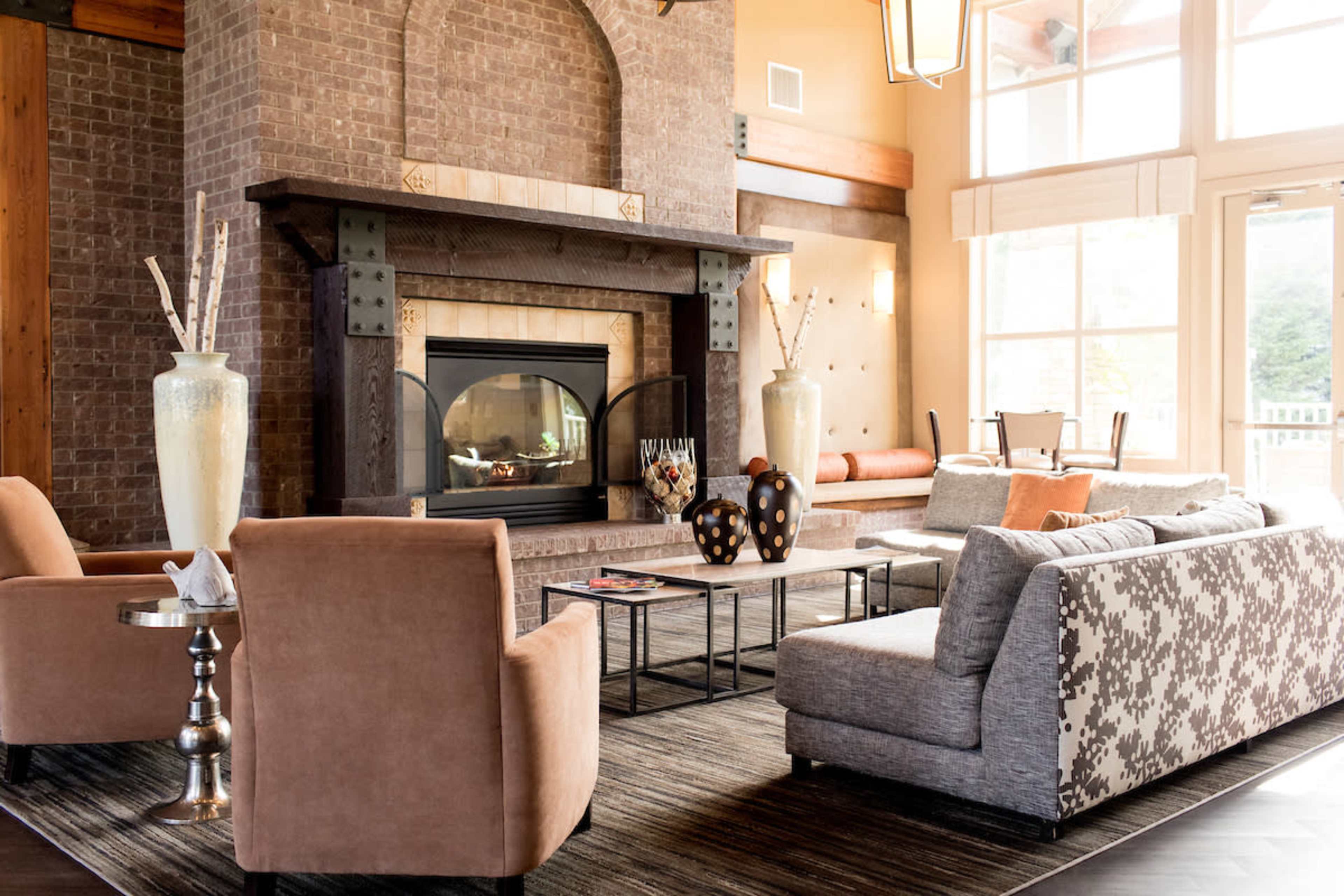 A spacious living room with a large brick fireplace, comfortable seating arrangements, and large windows allowing natural light to enter.