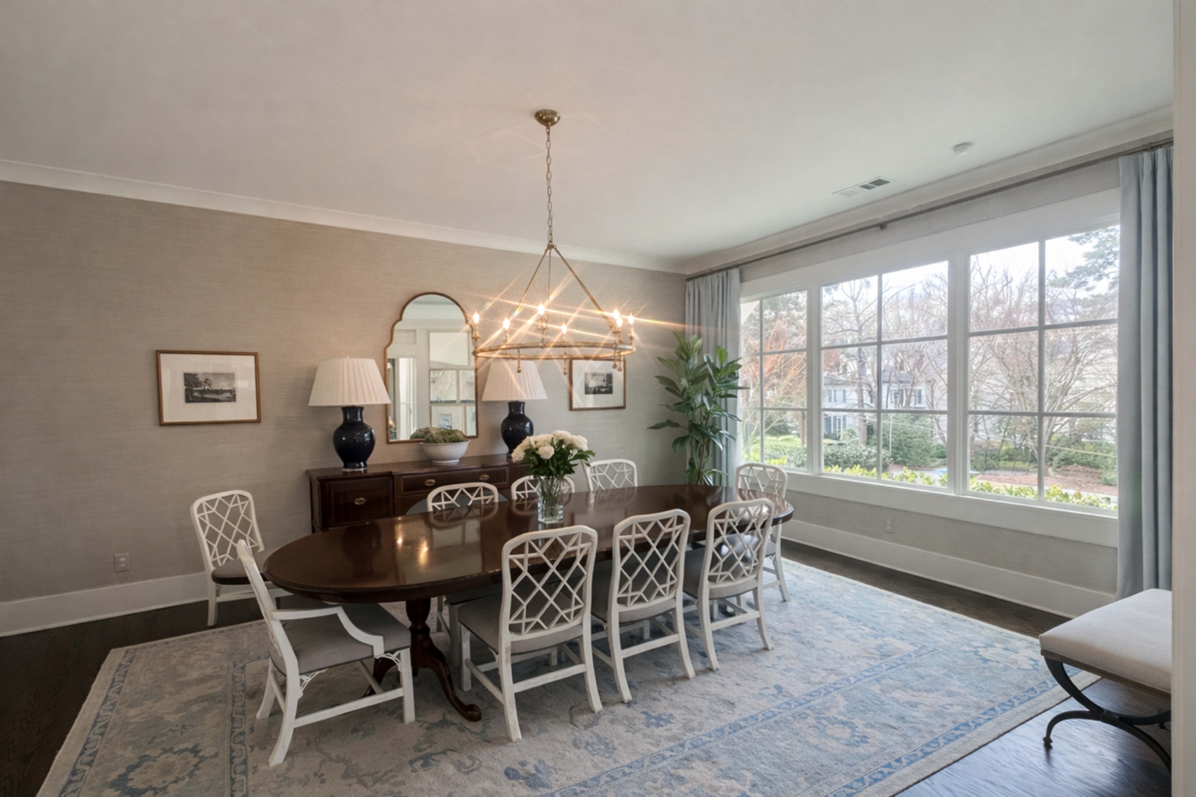 A dining room features a large oval table surrounded by white chairs, with a chandelier above and large windows offering natural light.