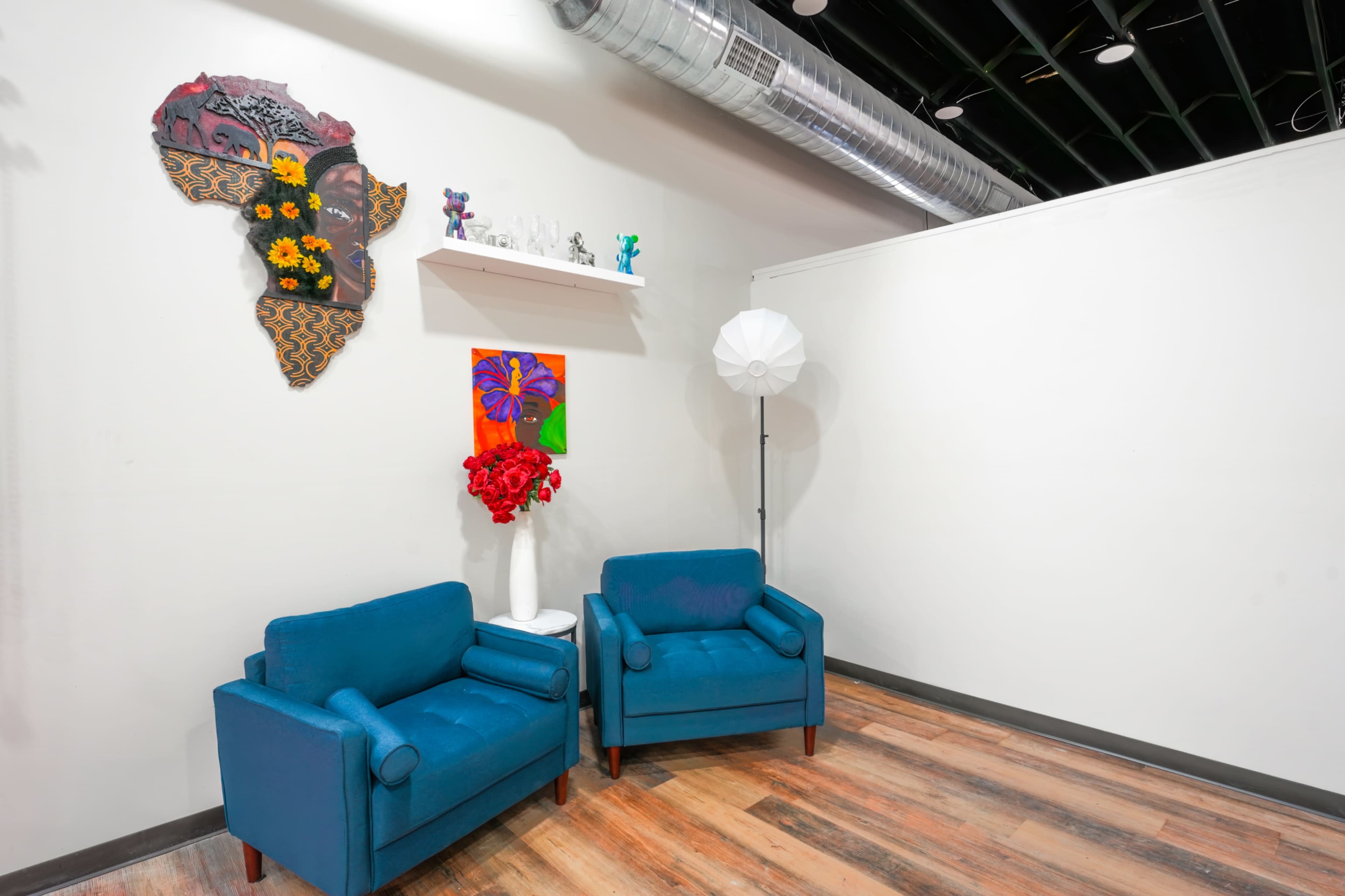 The image shows a well-lit corner of a room featuring two blue chairs, a white floor lamp, a vibrant wall decoration of Africa made from patterned fabric and flowers, and a shelf with small decorative items.