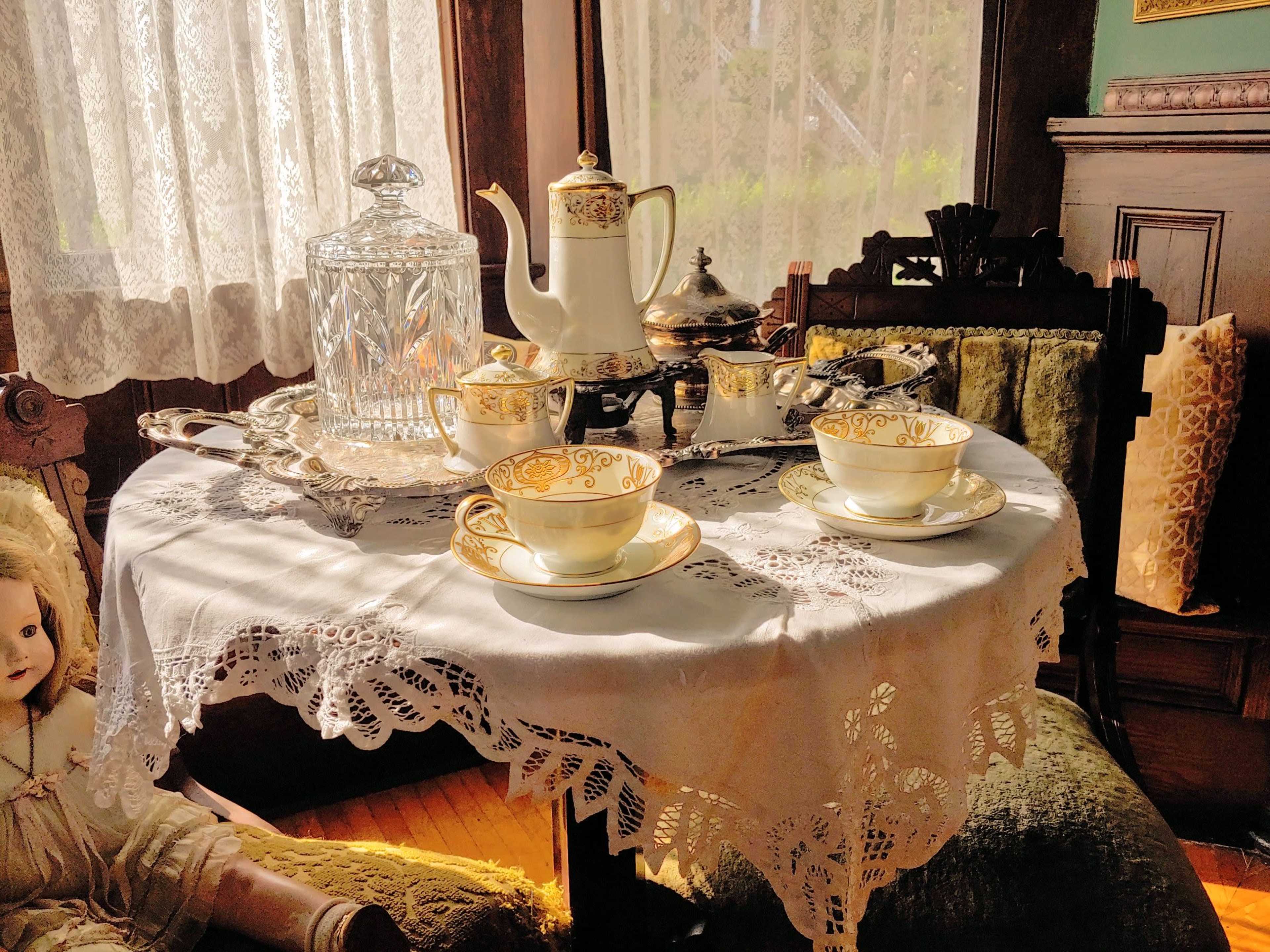 A polished tea set with ornate designs is arranged on a lace tablecloth beside a doll in a vintage room with sunlight streaming through a window.