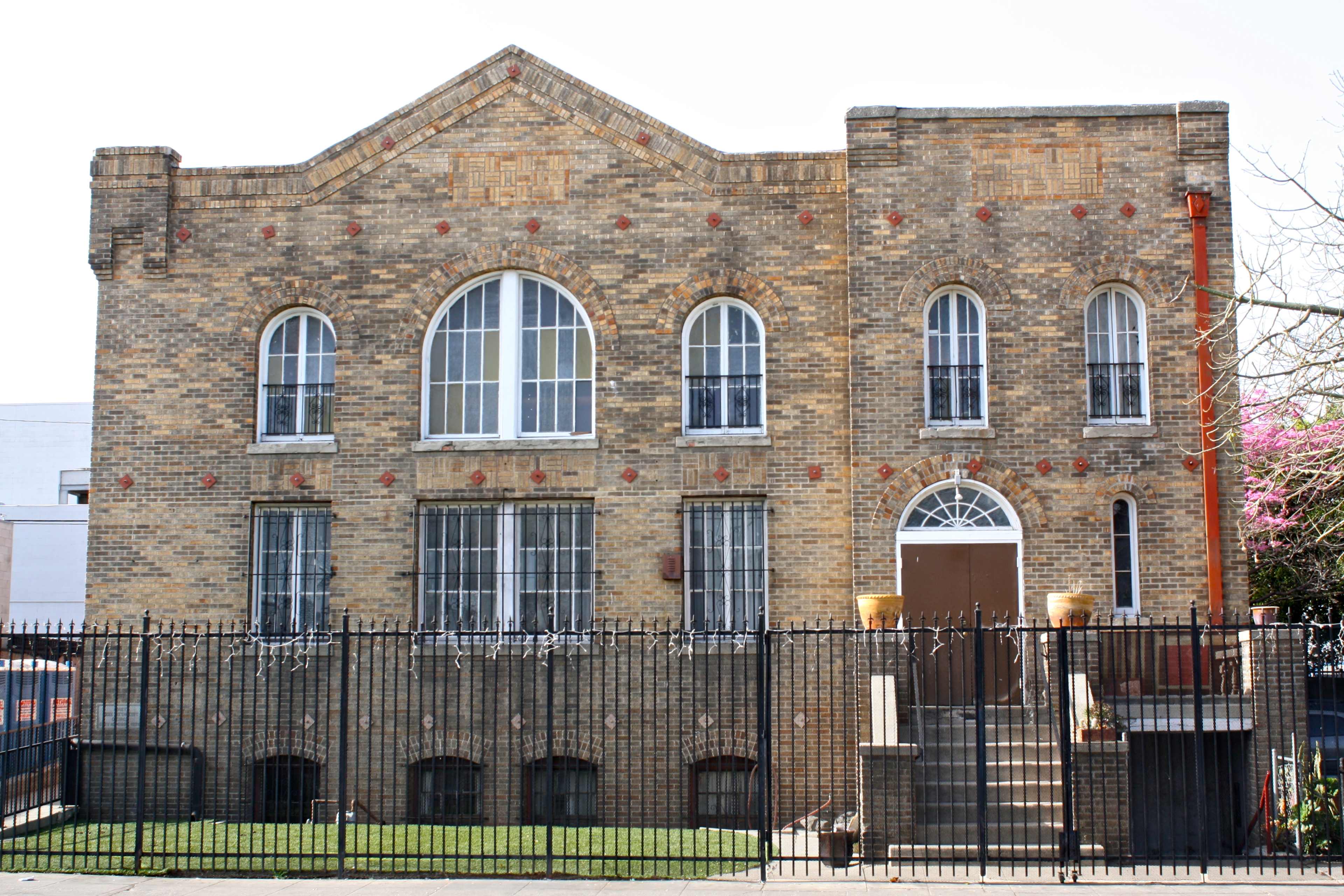A brick building with multiple arched windows and a gated entrance, featuring a staircase leading up to the main door.