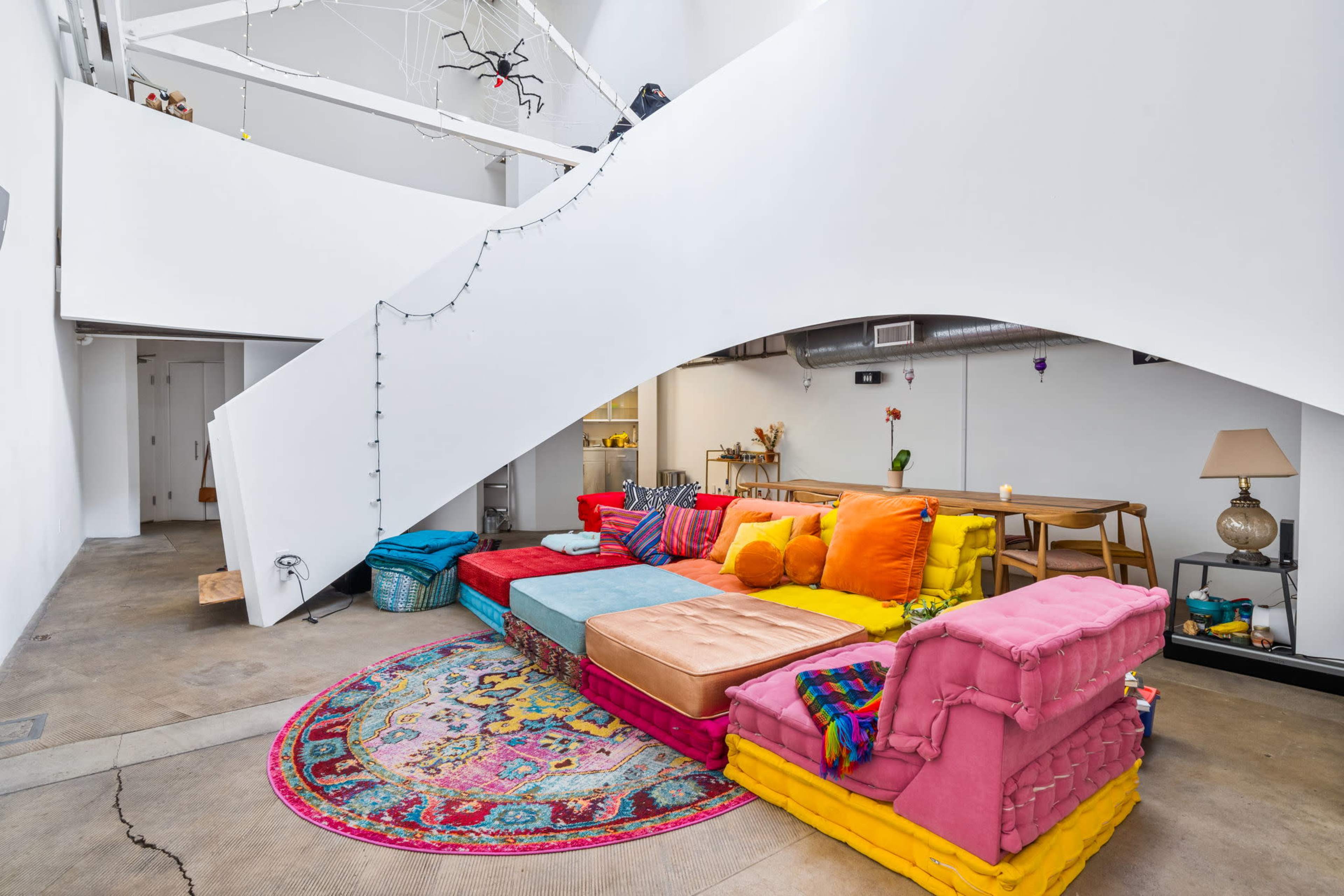 The image shows a modern interior space with a colorful seating area featuring vibrant cushions arranged around a decorative rug, beneath a sweeping white staircase.