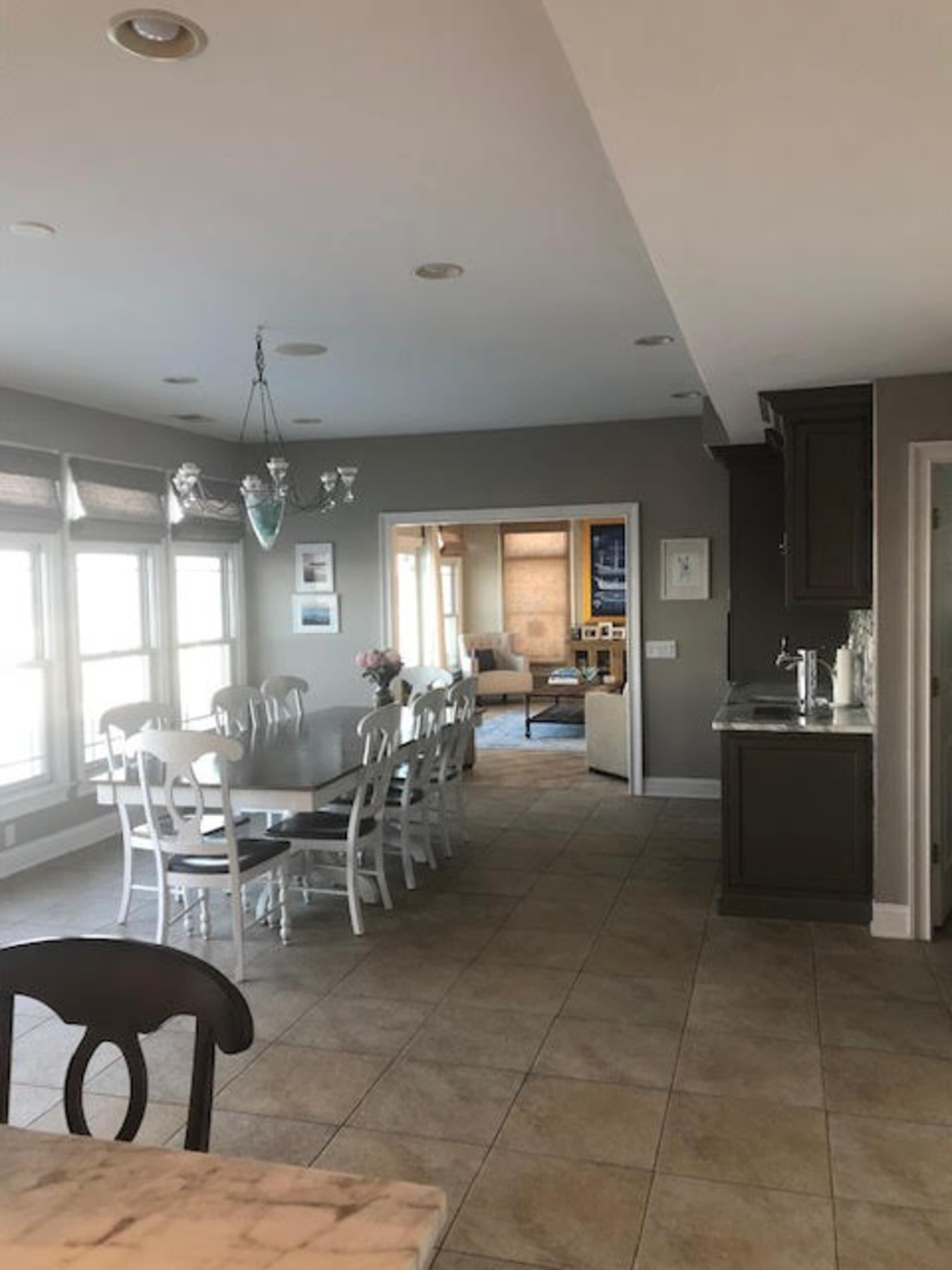 A spacious dining area with a long table and white chairs, leading into a living room visible through an open doorway.