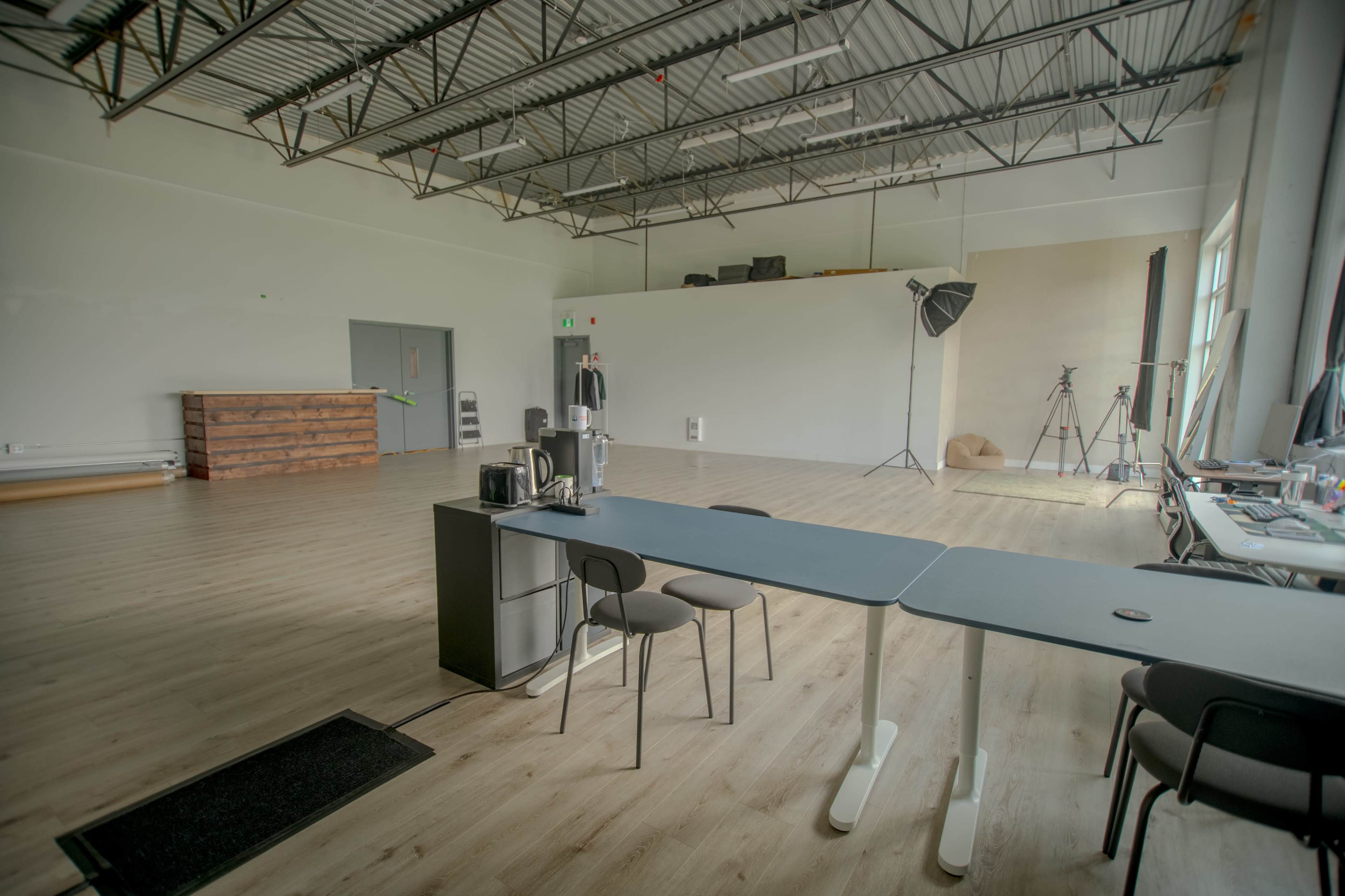 The image shows a spacious, empty studio with wooden flooring, a table with chairs, and photography equipment set up against the walls.
