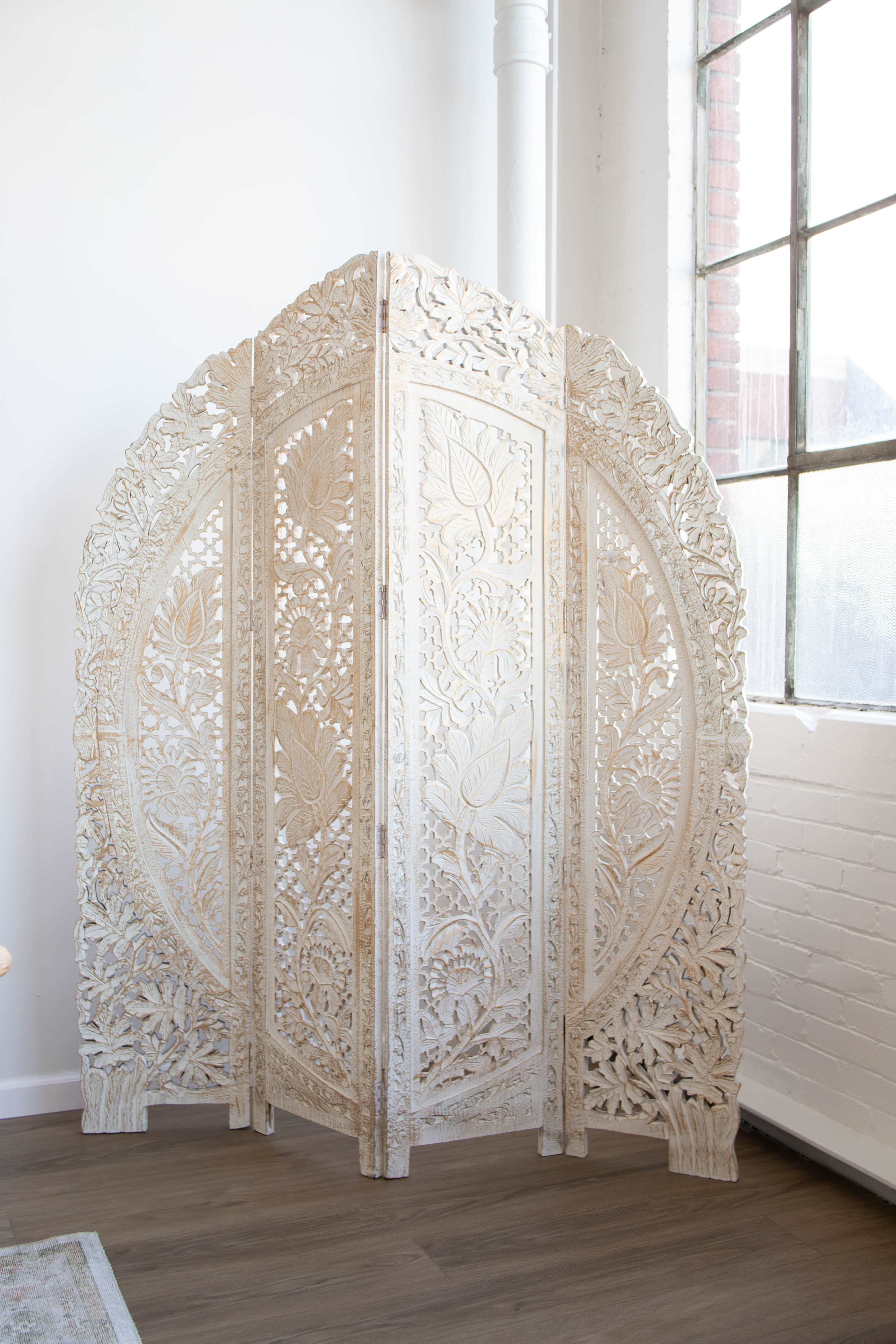 An intricately carved white wooden room divider with floral designs set against a wall near large windows.