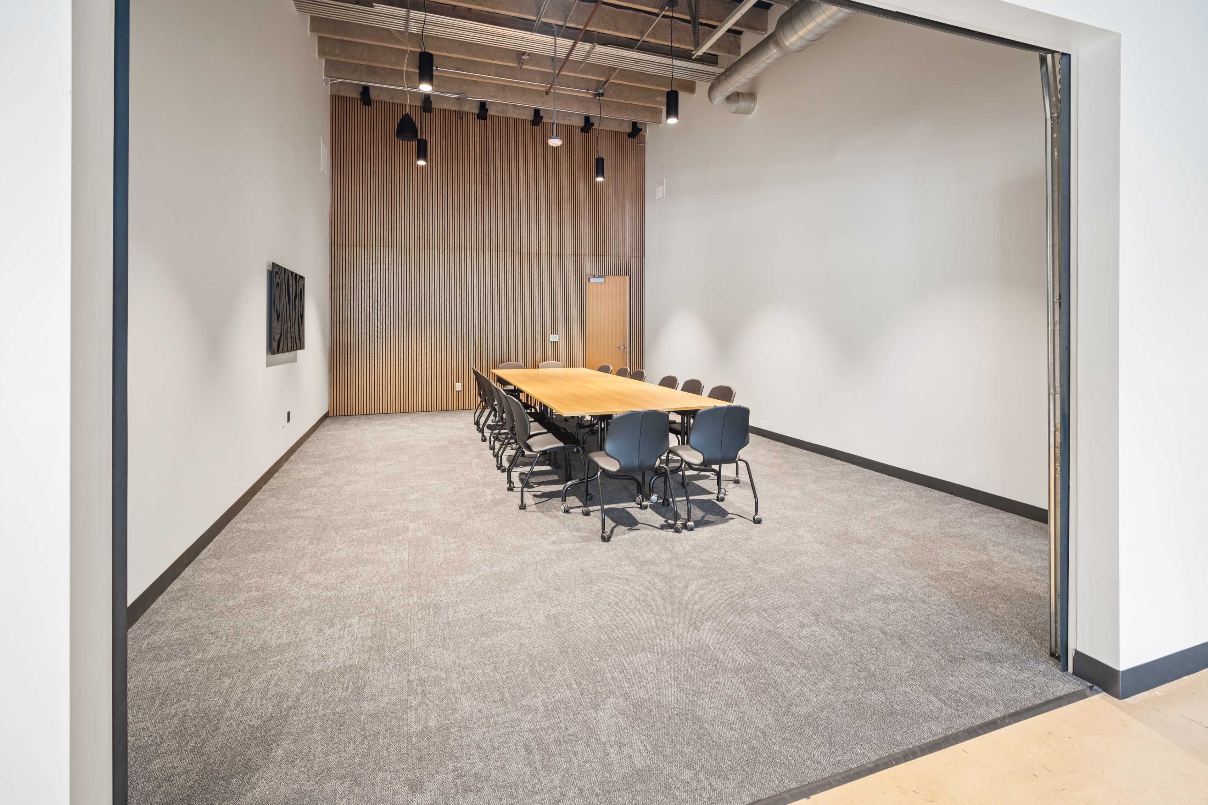 The image shows a spacious conference room with a long wooden table surrounded by black chairs, featuring a large blank wall and wood paneling in the background.