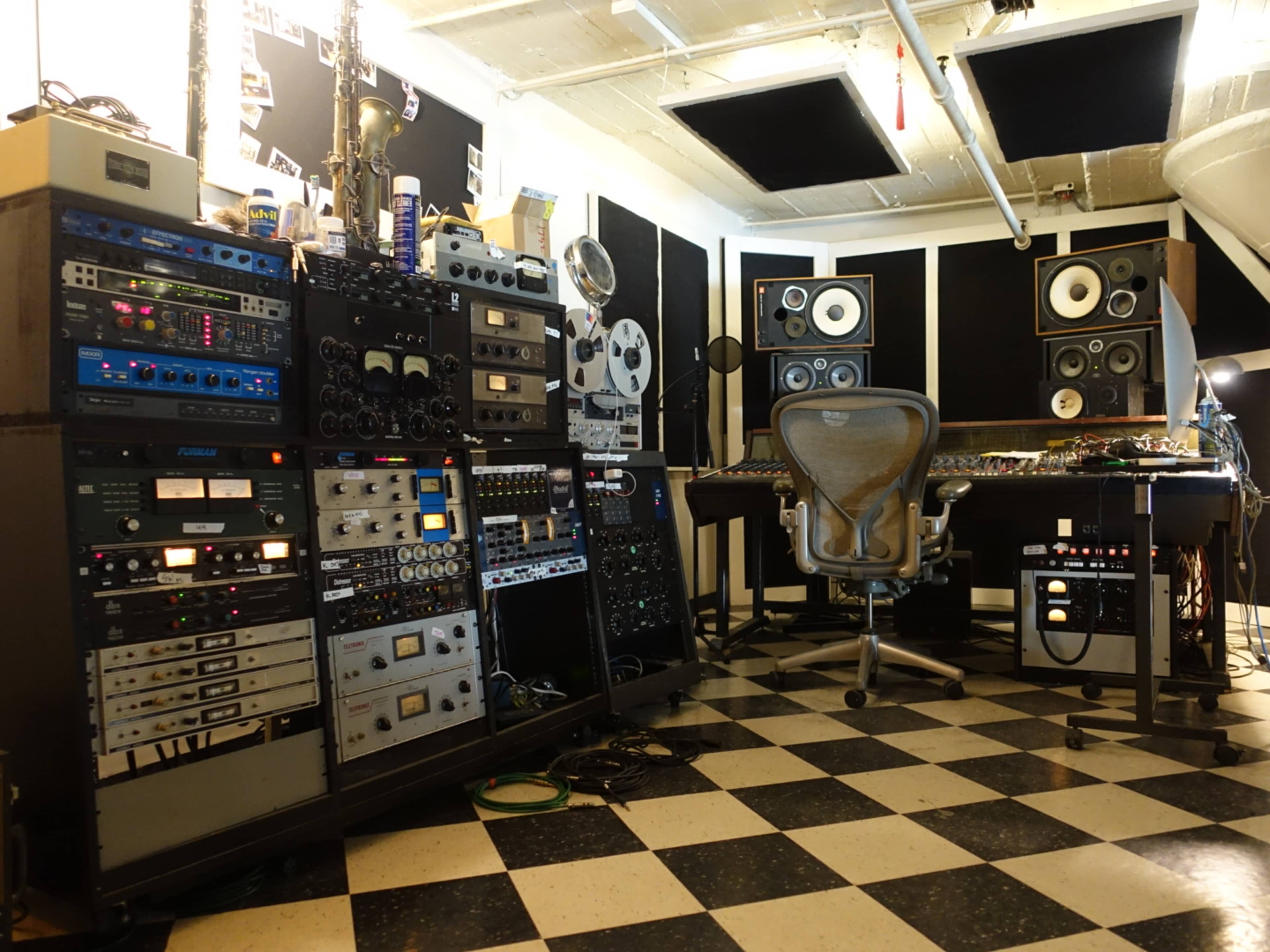 The image shows a music production studio with various audio equipment, including racks of signal processors and a mixing console, arranged alongside a chair and wall-mounted speakers.