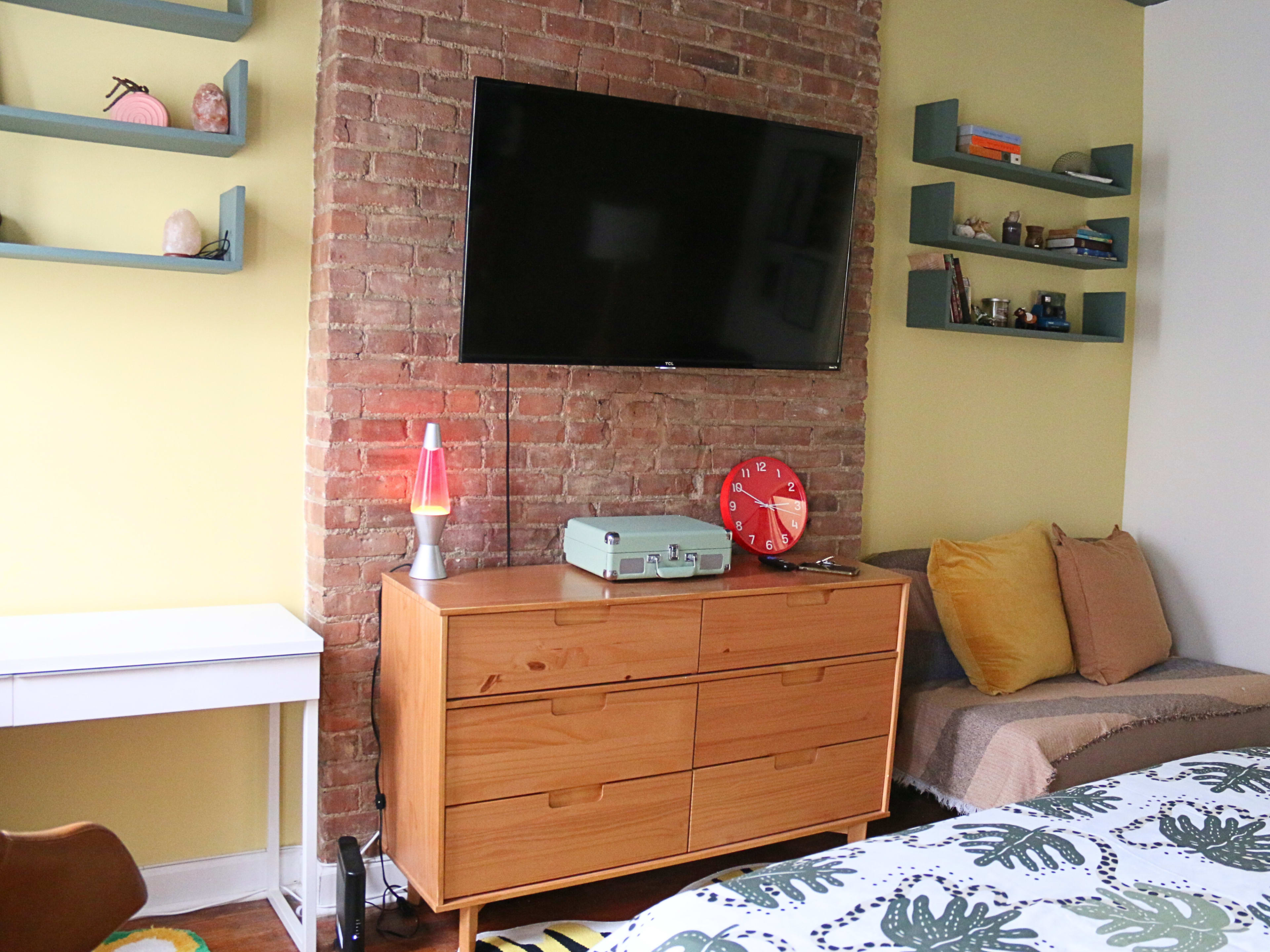 The image shows a cozy room featuring a brick wall with a flat-screen TV mounted on it, a wooden dresser, and a seating area with decorative items on shelves.
