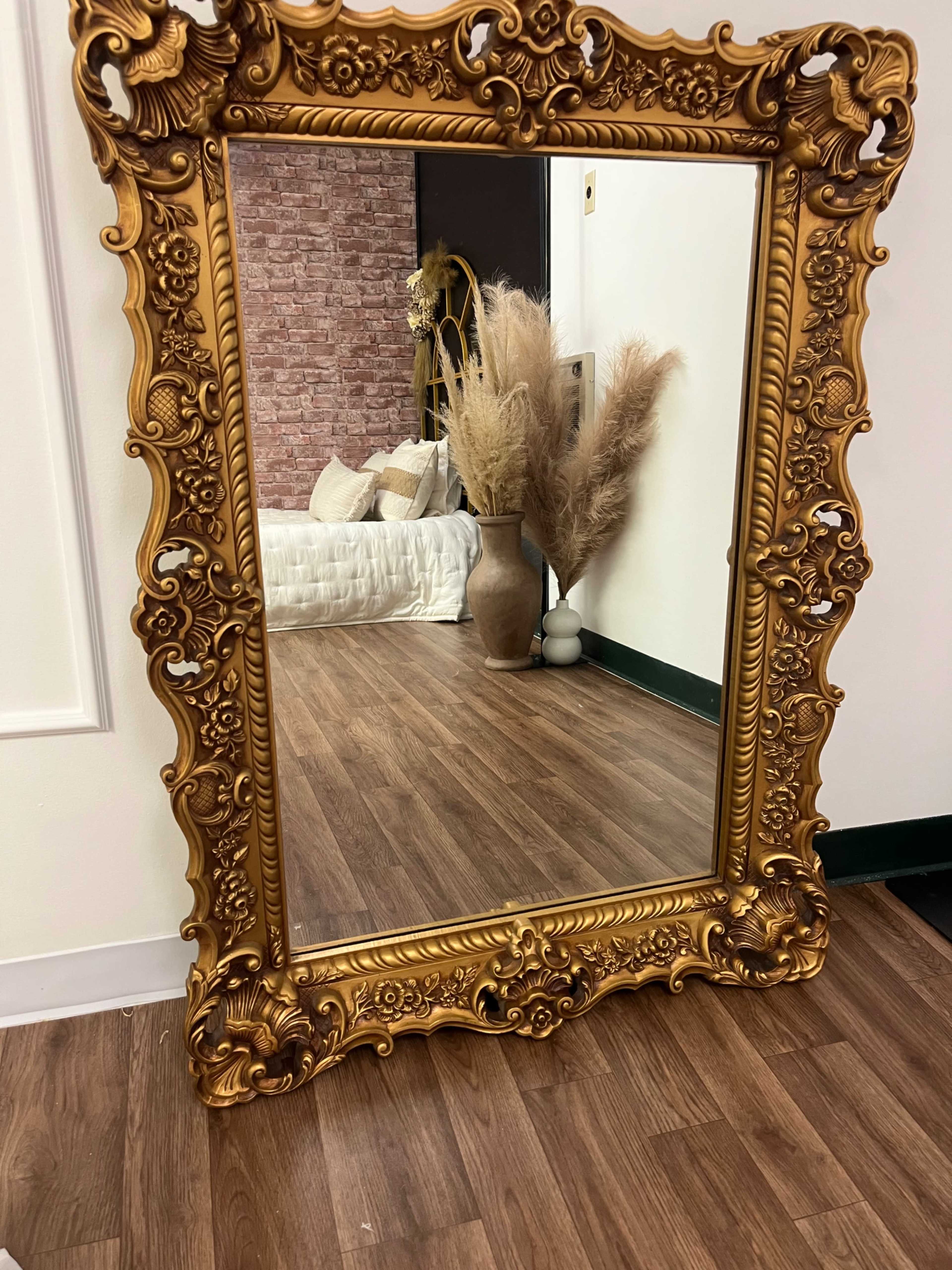 A large ornate gold mirror stands against a wall, reflecting a room with a brick accent wall, a vase, and decorative plants.
