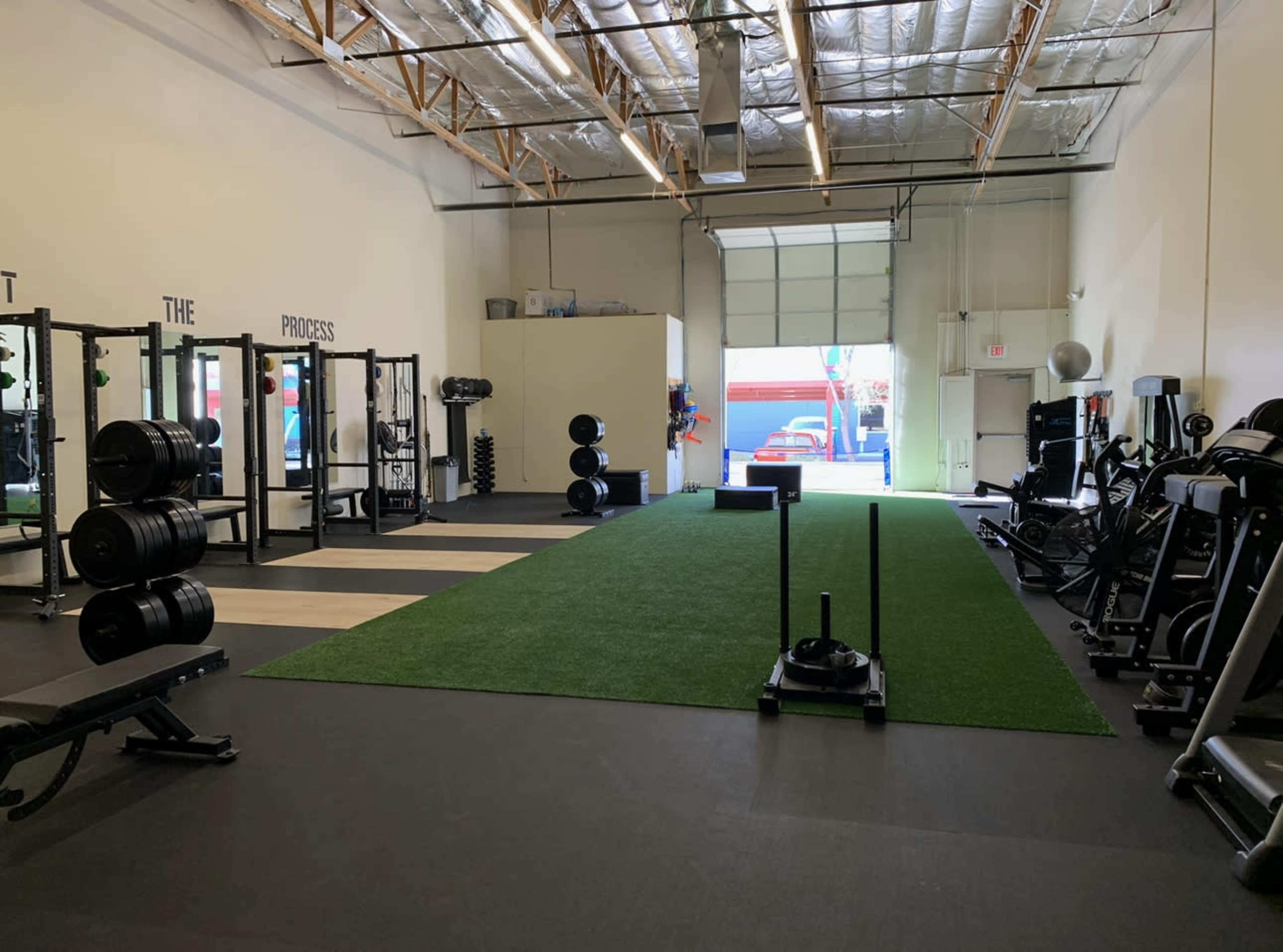 A fitness facility with a turf area, various weightlifting equipment, and an open garage door leading outside.