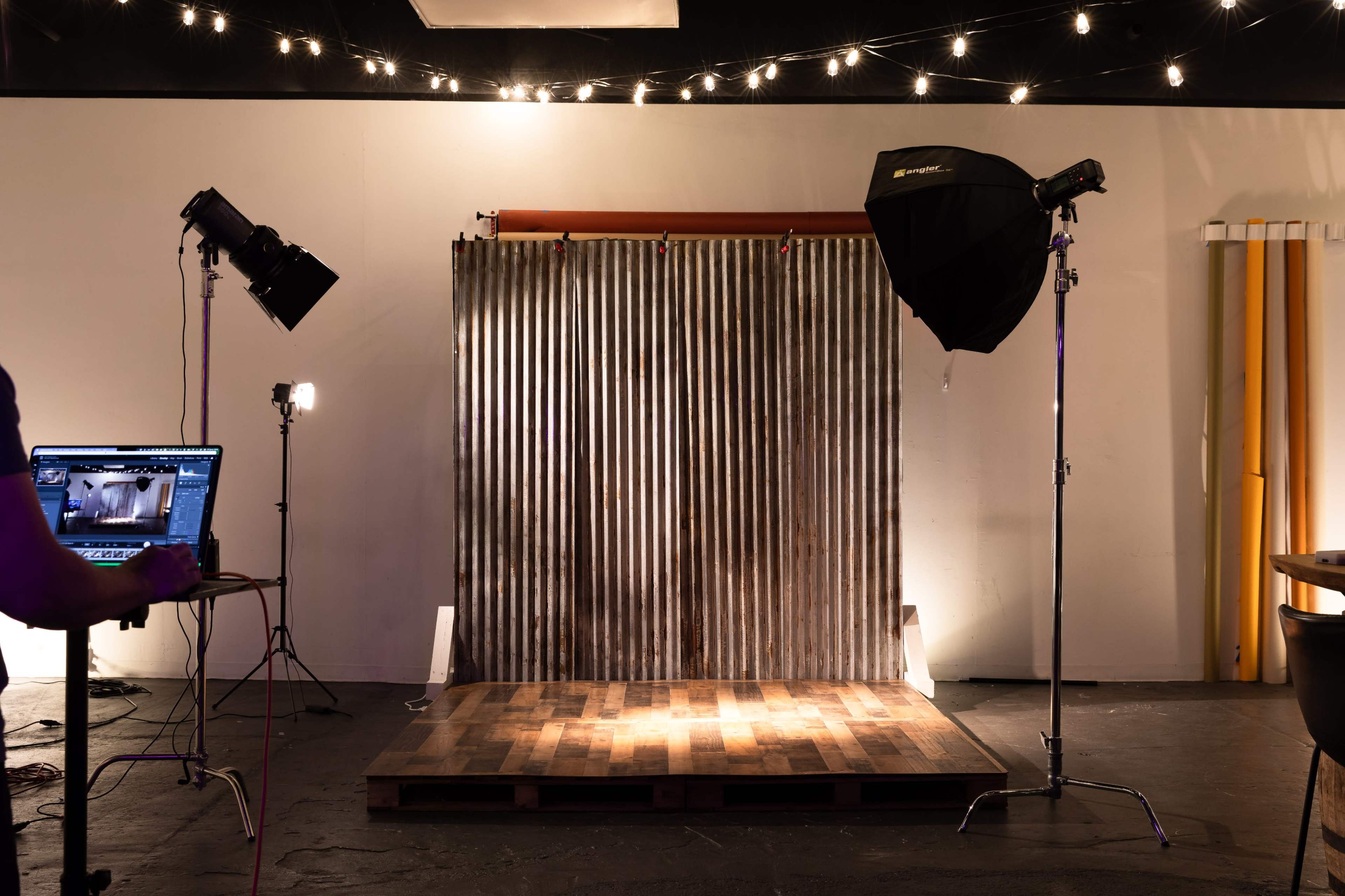 The image shows a photography studio setup with a corrugated metal backdrop, wooden flooring, and several lighting equipment positioned around the area.