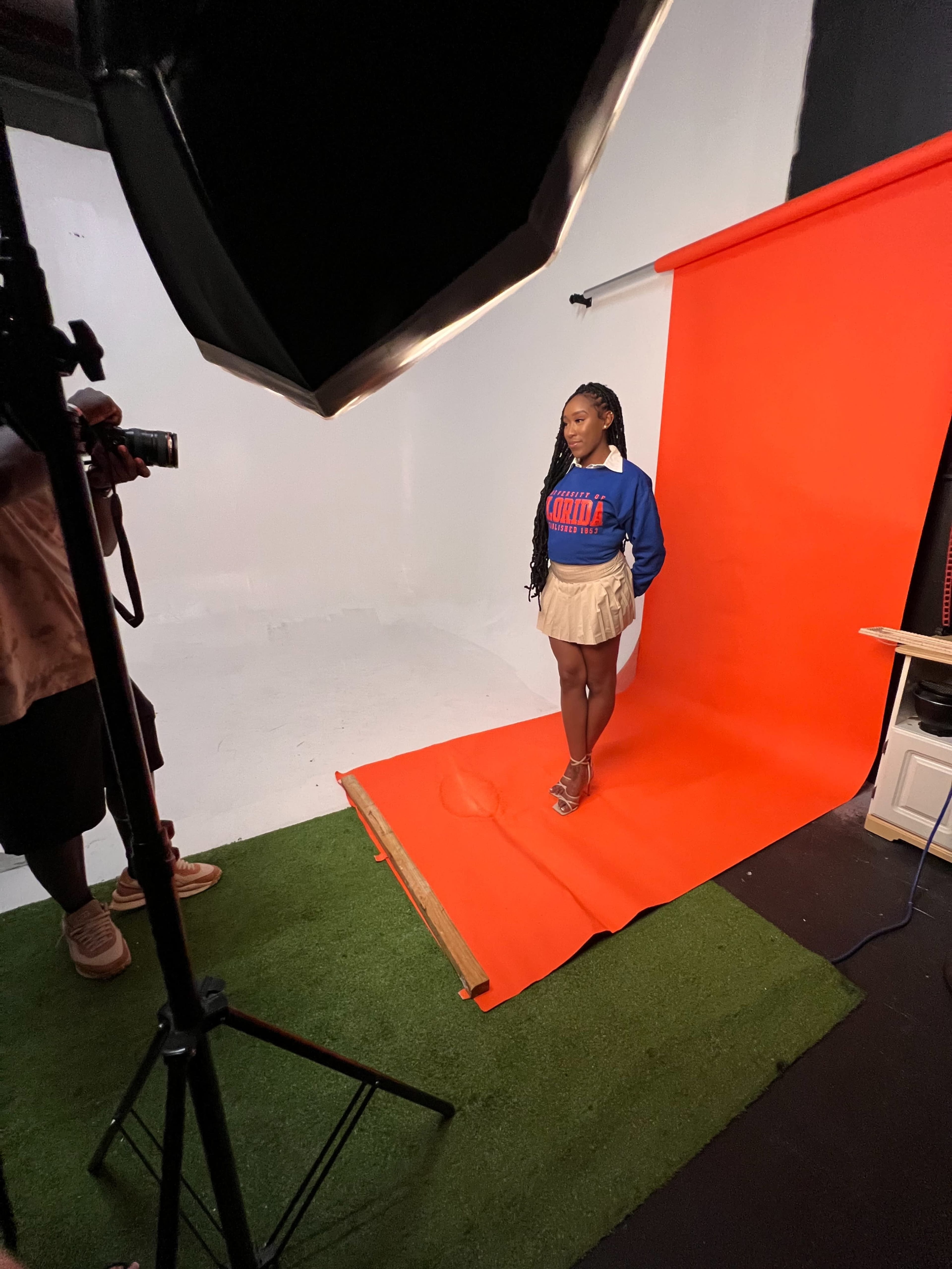 A person stands on a red backdrop in a photo studio while another individual prepares to take their picture.