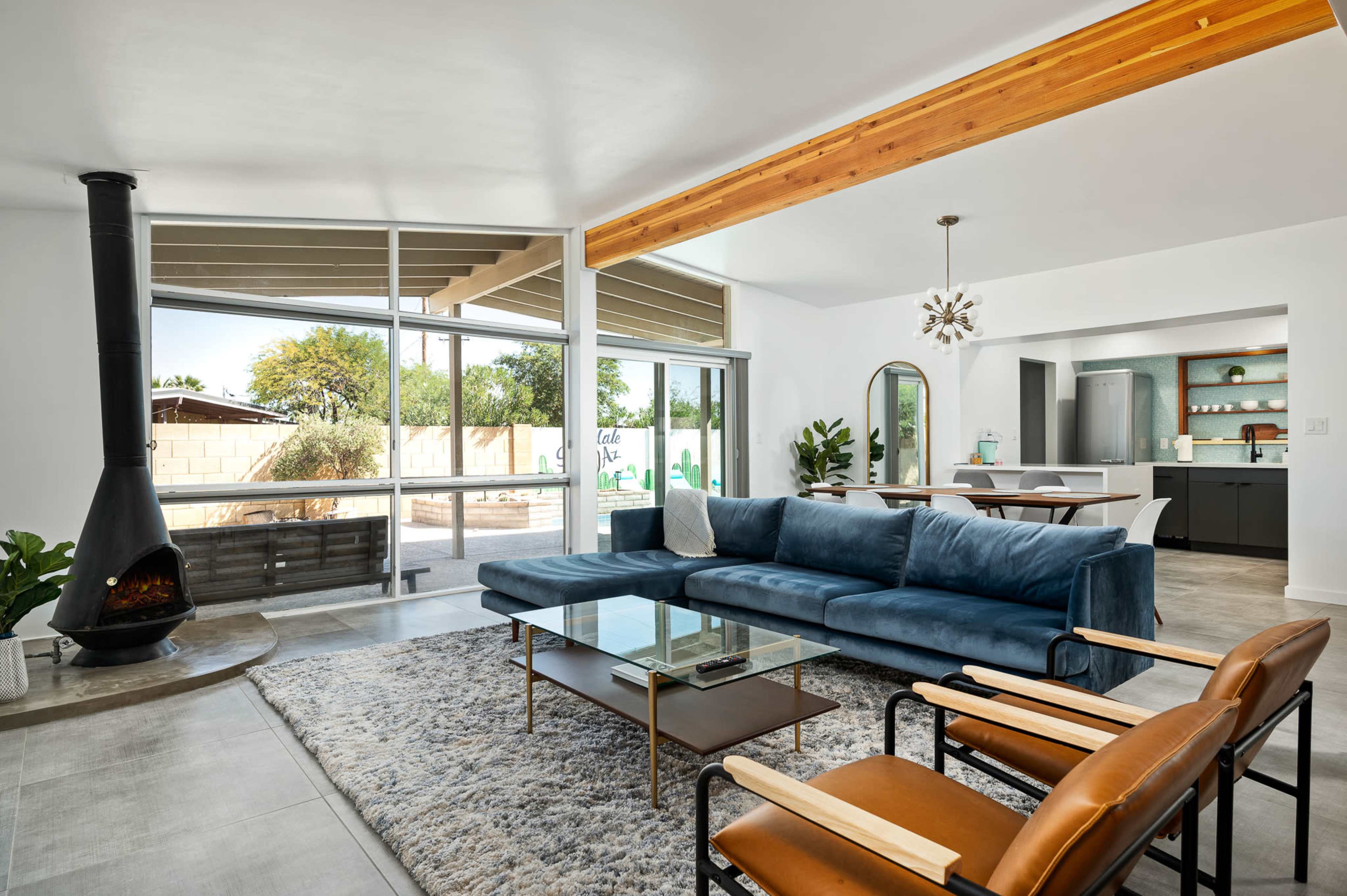 A modern living room featuring a blue sectional sofa, a glass coffee table, and a unique fireplace, with large windows overlooking a backyard.