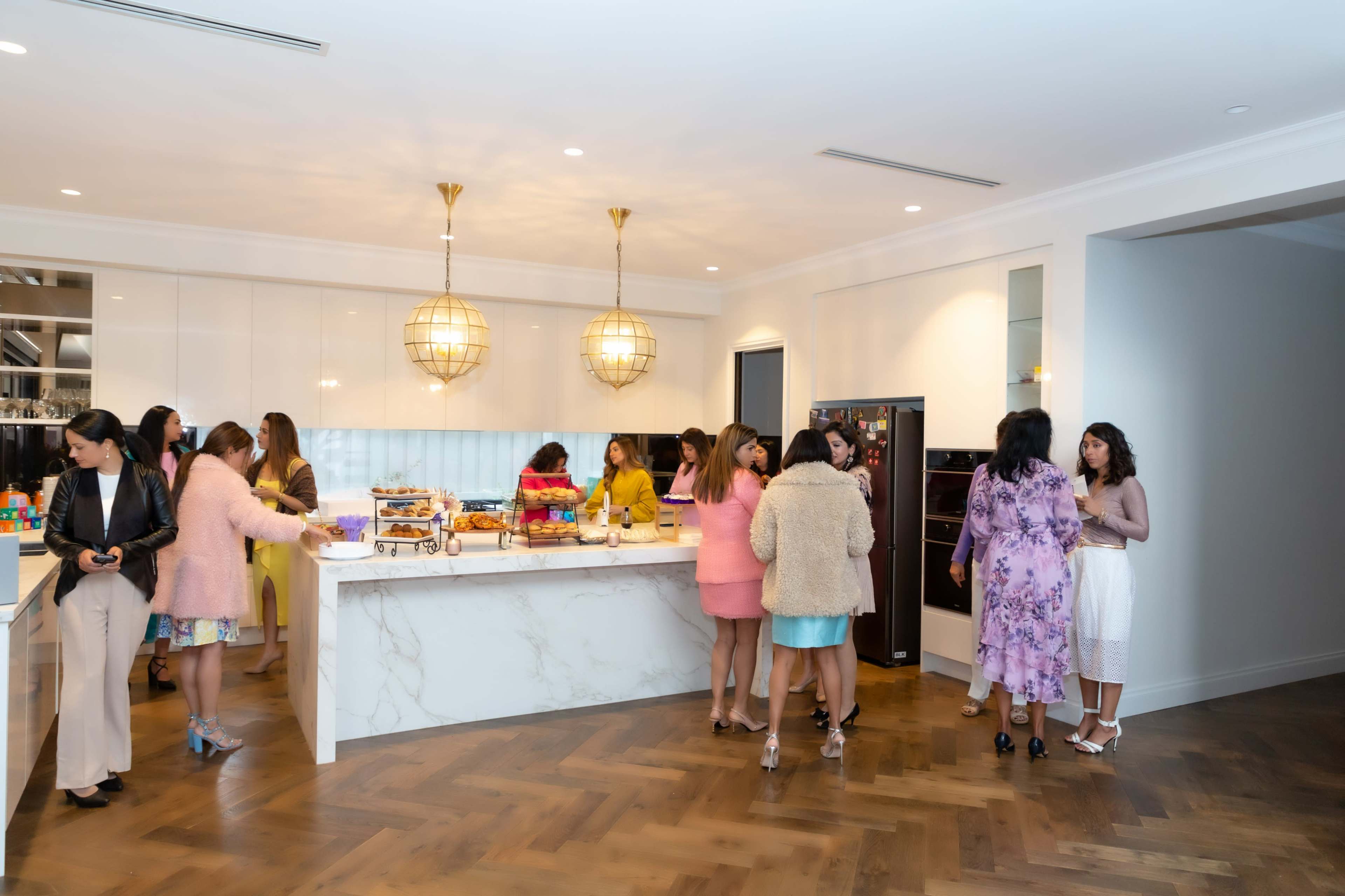 A group of women gathers in a modern kitchen with a large marble island, preparing food and drinks.