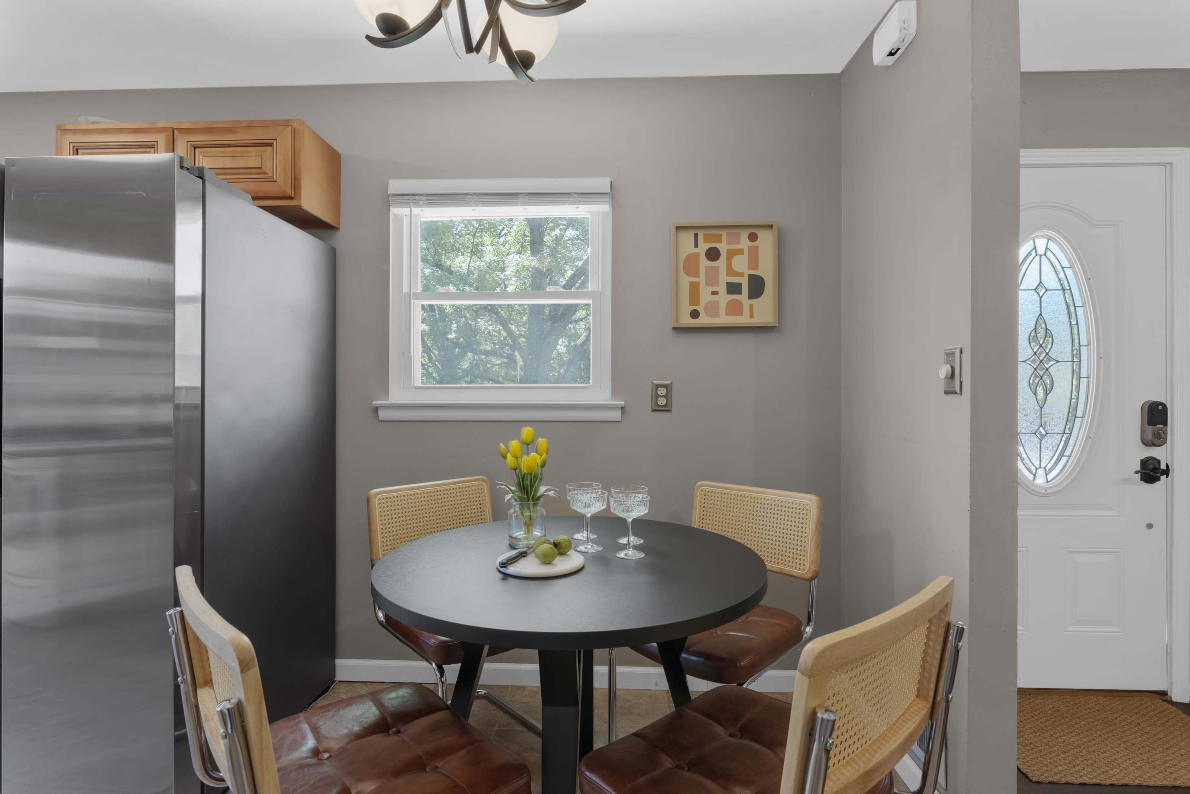 A small dining area features a round black table set with glasses and fruit, surrounded by four chairs, next to a modern refrigerator and a front door with decorative glass.