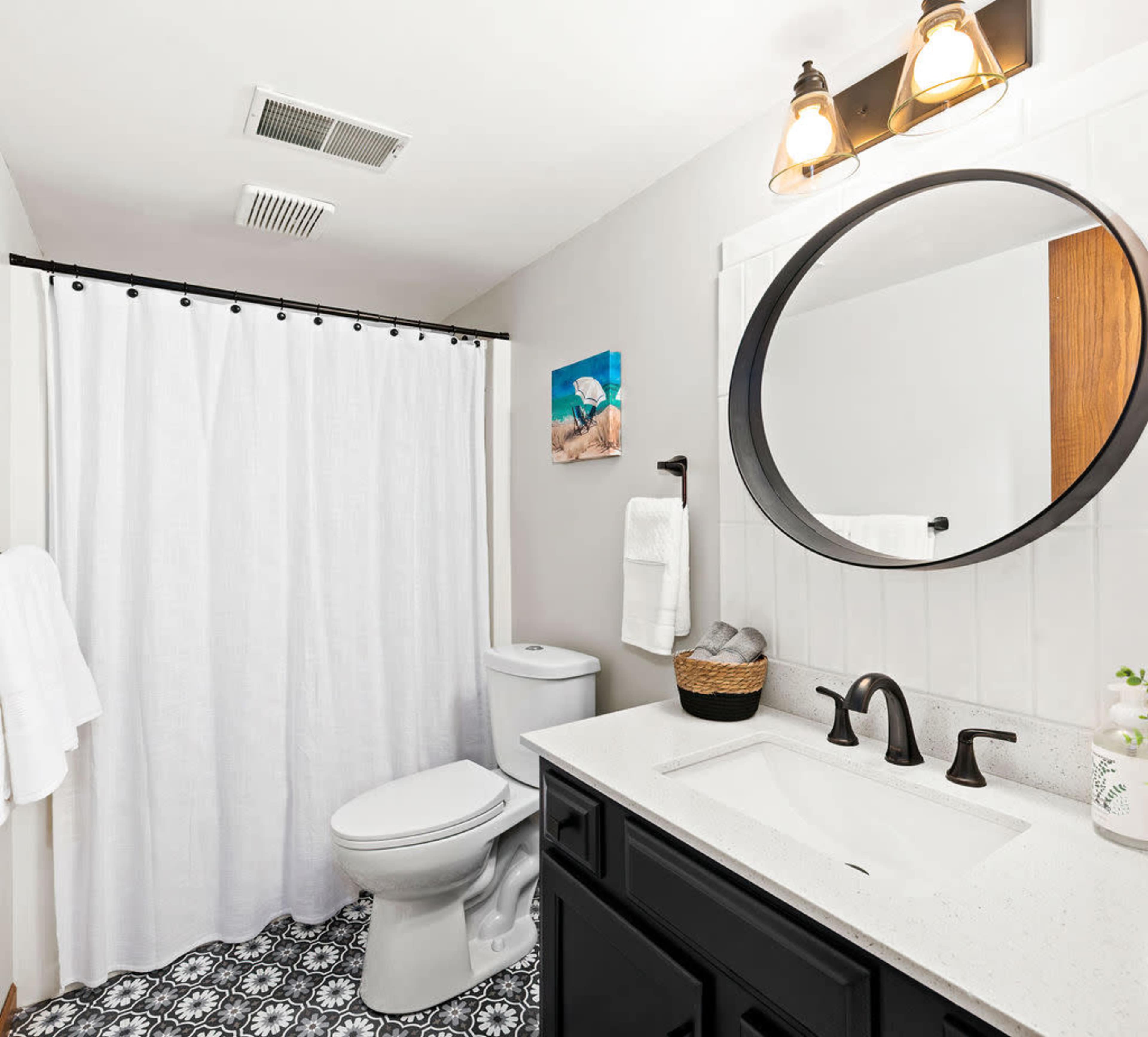 The bathroom features a white shower curtain, a round mirror above a sink, a modern toilet, and patterned tile flooring.