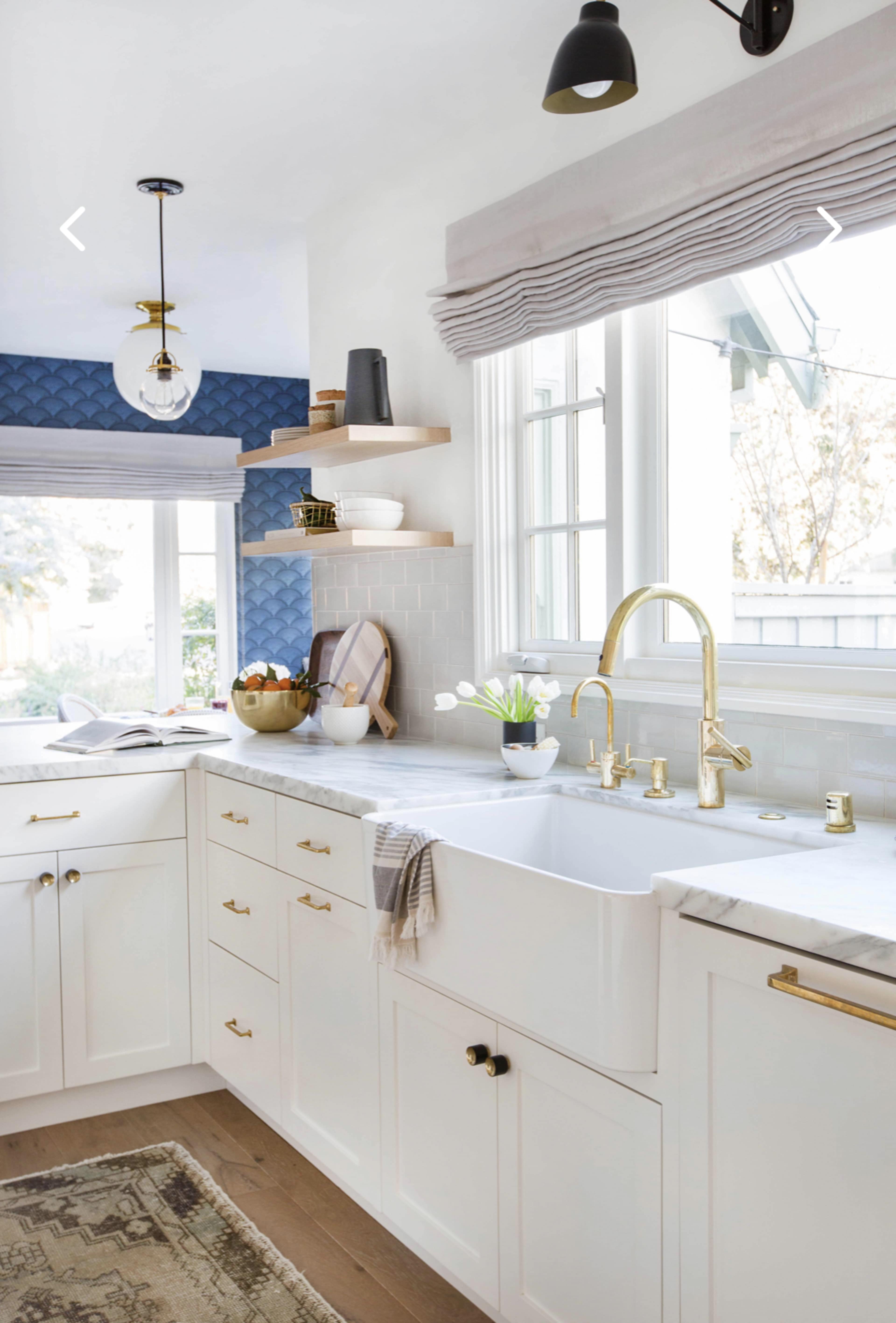 A modern kitchen with white cabinetry, a farmhouse sink, and brass fixtures, complemented by blue wall accents and natural light from windows.