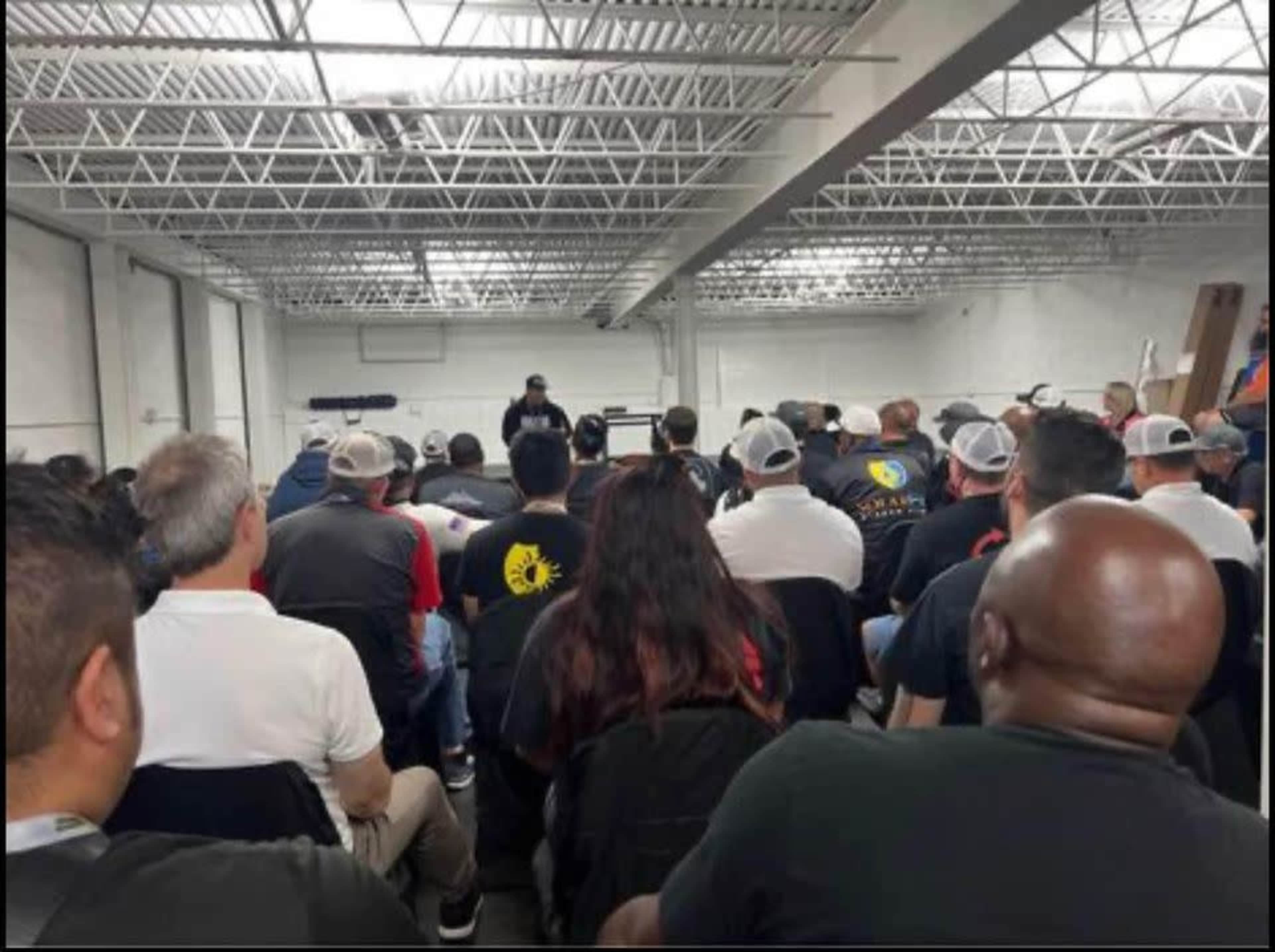 A crowd of attendees sits in chairs facing a speaker at the front of a large, well-lit room.