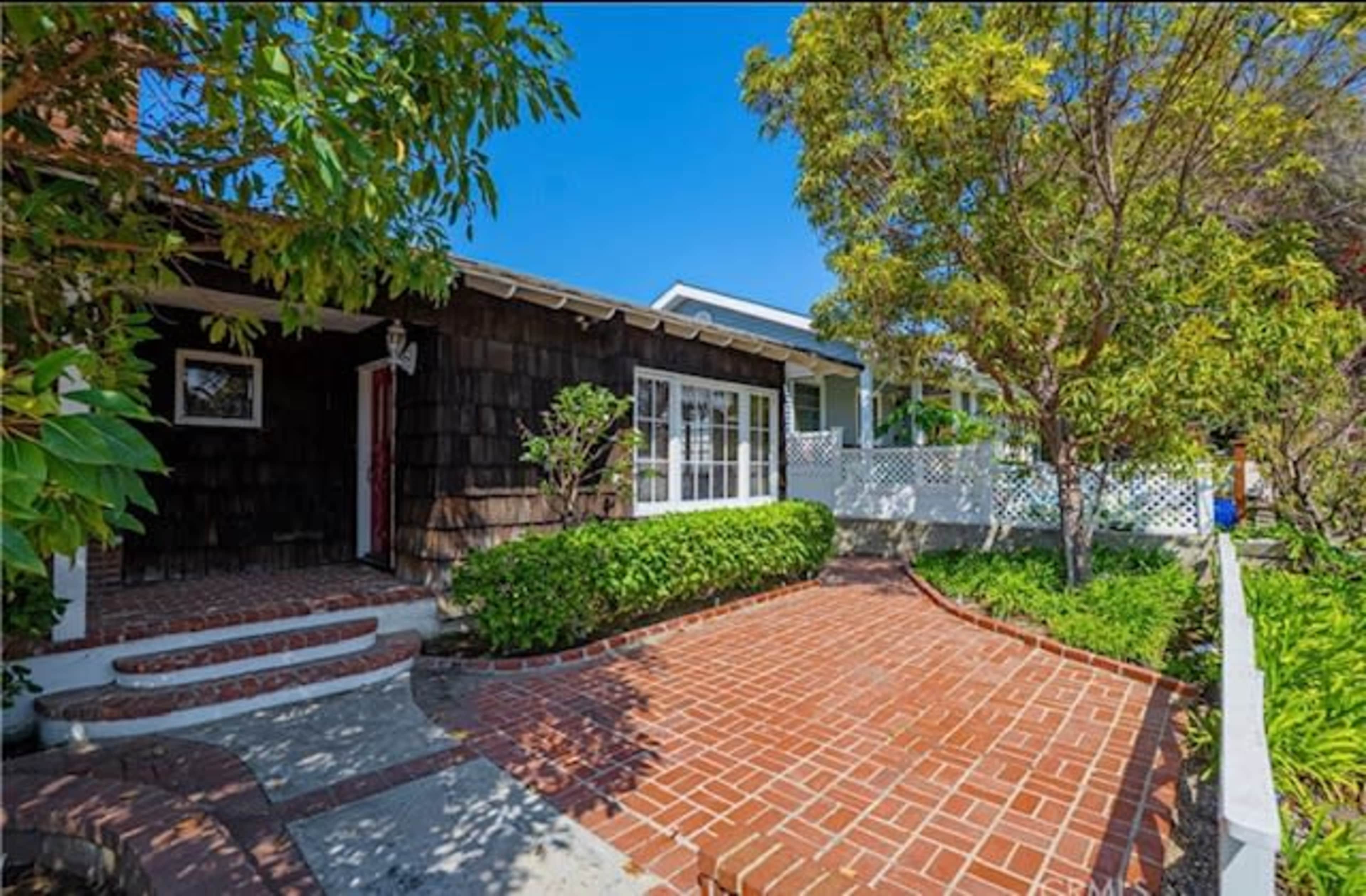 The image shows a brick pathway leading to a single-story house with wooden siding and a front porch, surrounded by various green plants and trees.