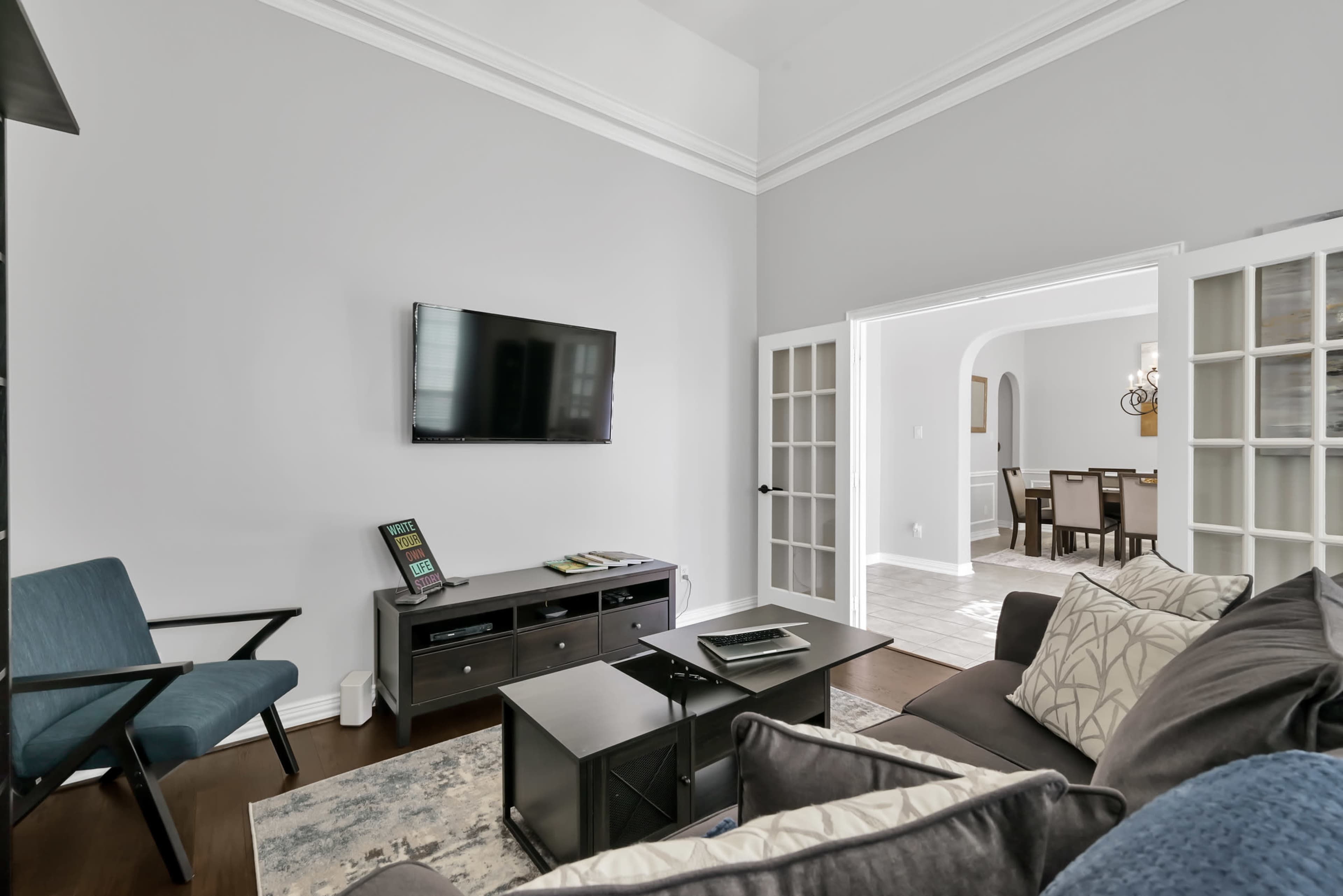 A modern living room features a mounted television, a black coffee table, and an armchair, with a view of a dining room through a set of glass-paned doors.