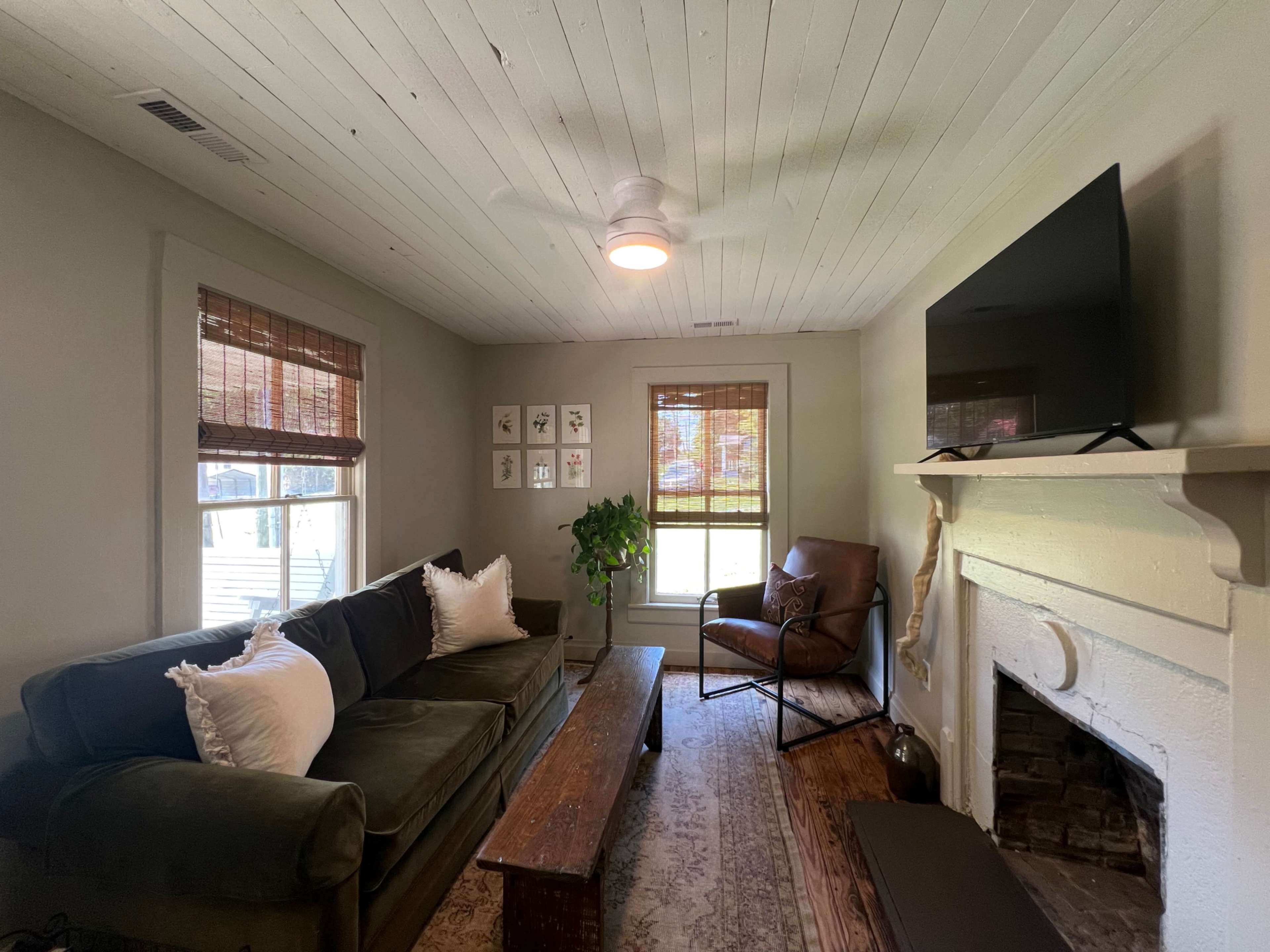 The image depicts a cozy living room featuring a dark green couch, a wooden coffee table, a brown armchair, and a mounted television above a white fireplace.