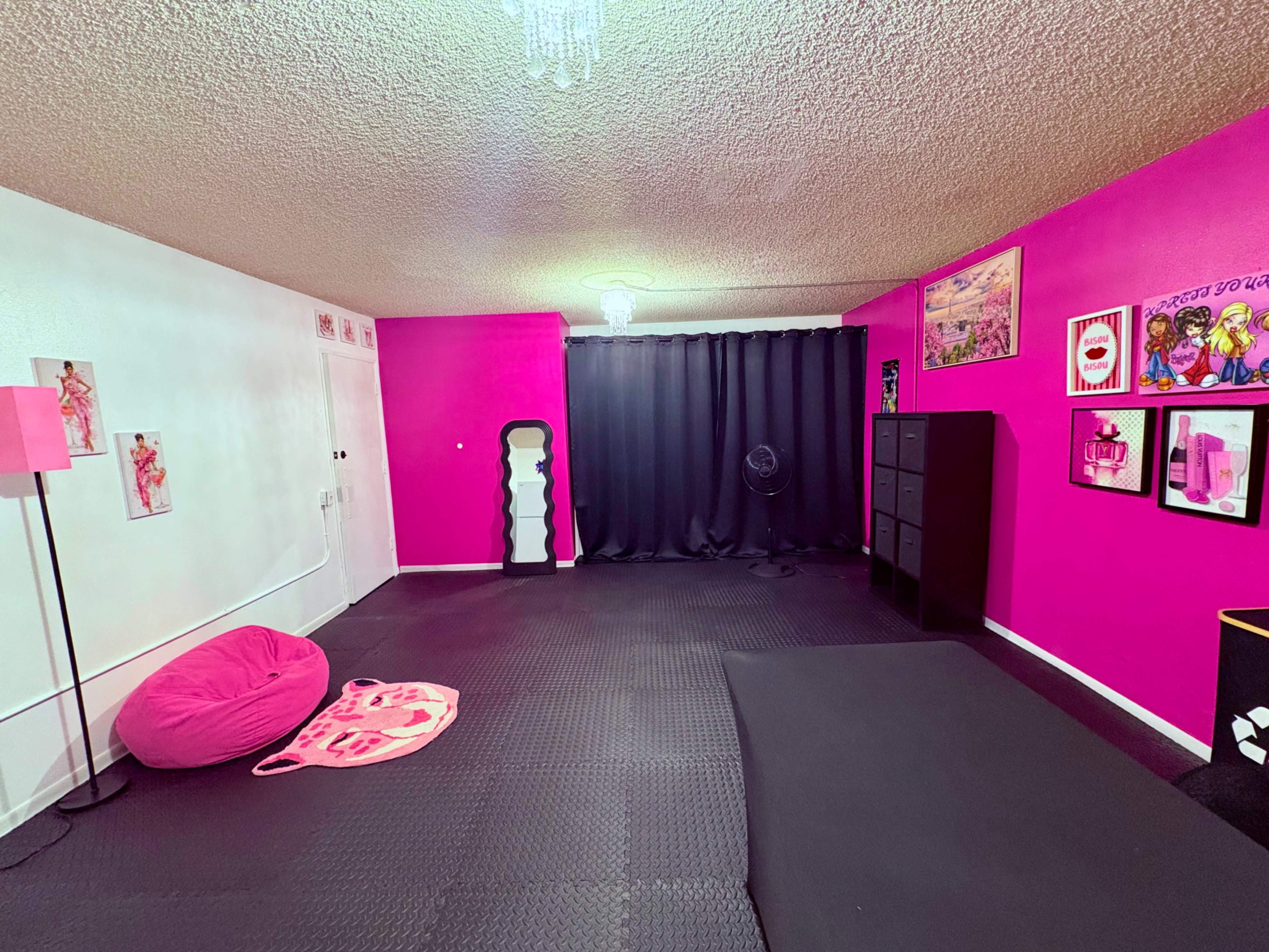 The room features bright pink walls, black curtains, a fan, a clothes rack, and a black storage unit, with a pink bean bag and a patterned mat on the floor.